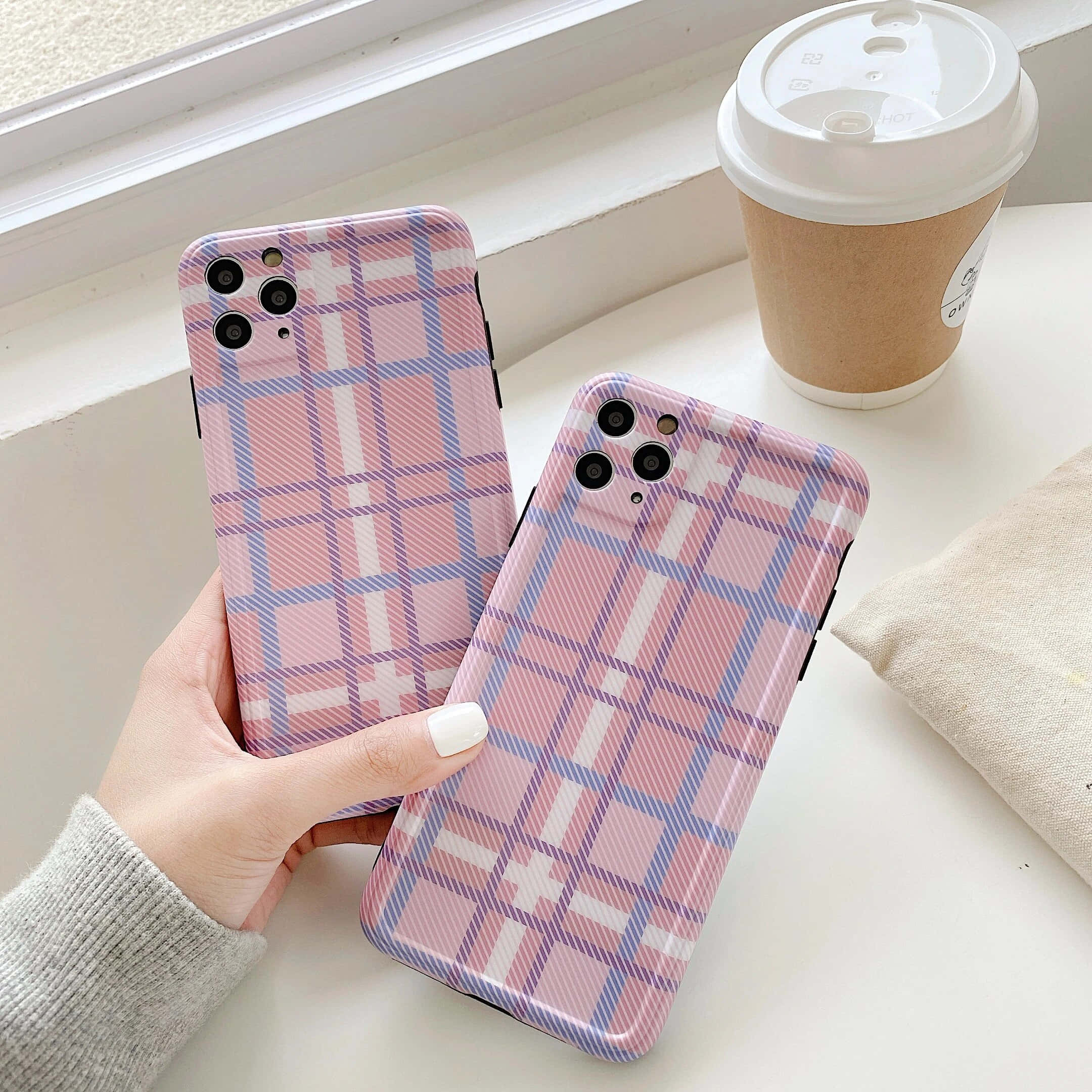 可愛 iPhone 桌布 桌布 壁紙 — 一隻手拿著兩個粉紅、紫和白格紋圖案的手機殼，放在窗台上的咖啡杯旁邊。