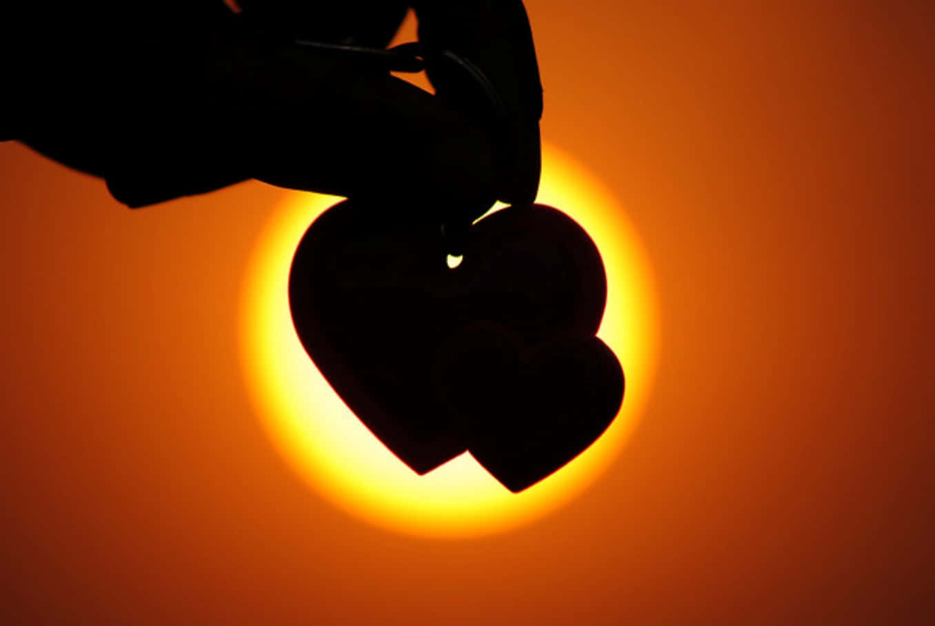 Heart Silhouette 桌布 壁紙 — 一隻手拿著兩個重疊的心形剪影，襯著發光的橙色日落。