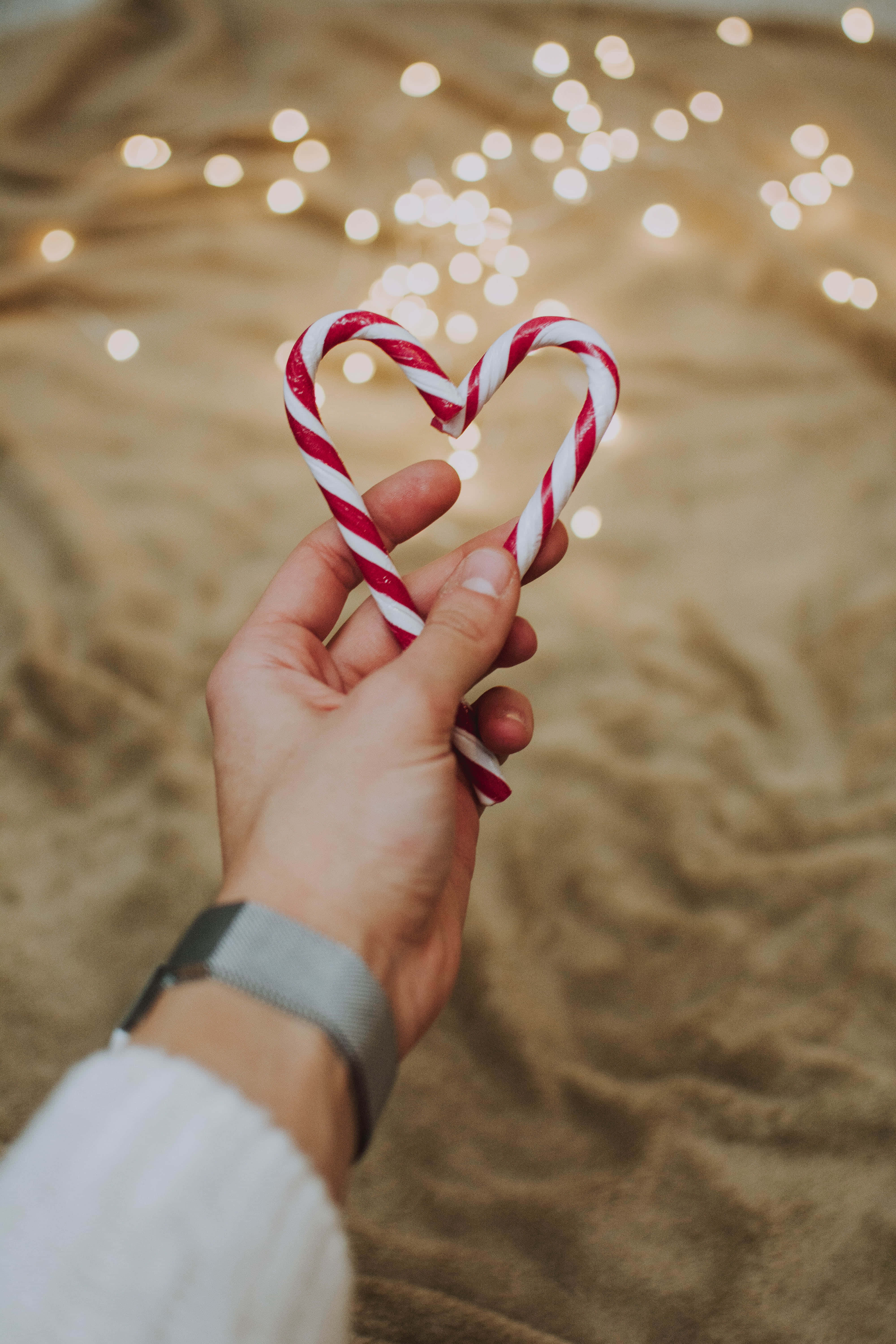 Christmas Scene 桌布 壁紙 — 一隻手拿著兩根紅白條紋的拐杖糖，排列成心形，背景是柔和散景的閃爍燈光。
