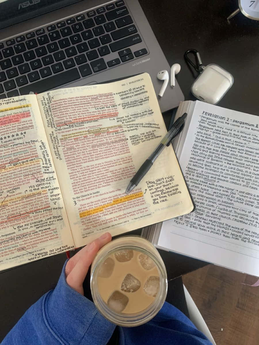 書桌 桌布 壁紙 — 一隻手拿著冰咖啡，旁邊放著打開的聖經和筆記型電腦，在木桌上，還有耳塞和筆。