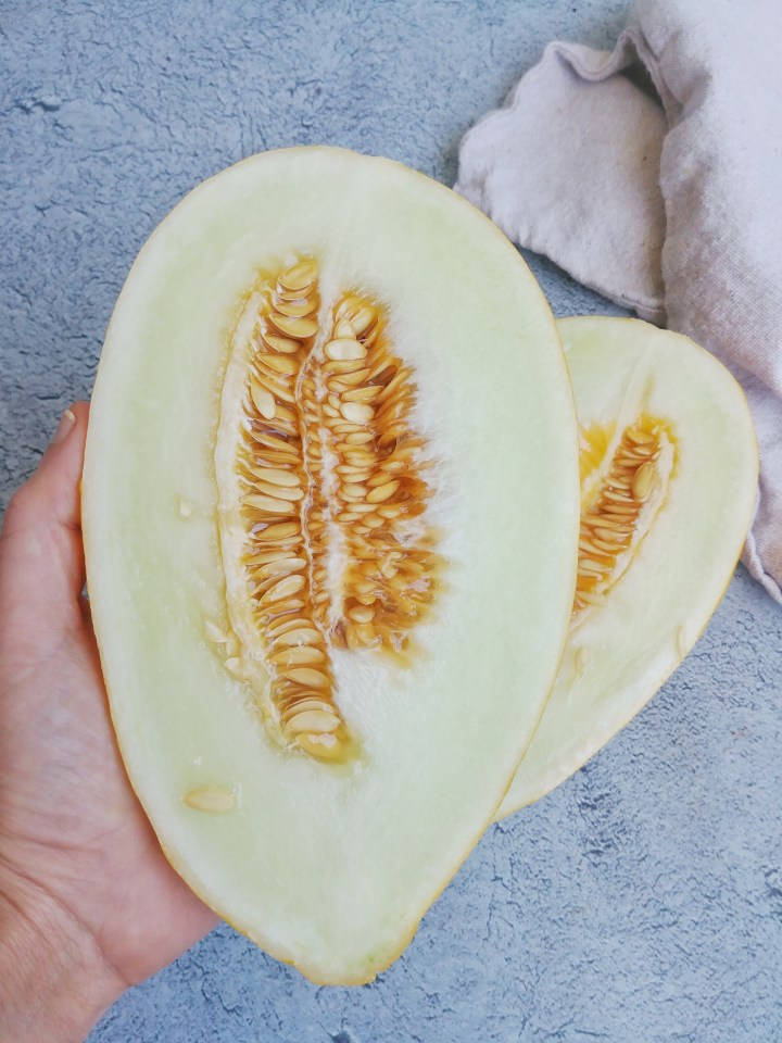 自然 桌布 壁紙 — 一隻手拿著半個哈密瓜，露出多汁的淡色果肉和琥珀色的種子，背景是帶紋理的藍色表面。