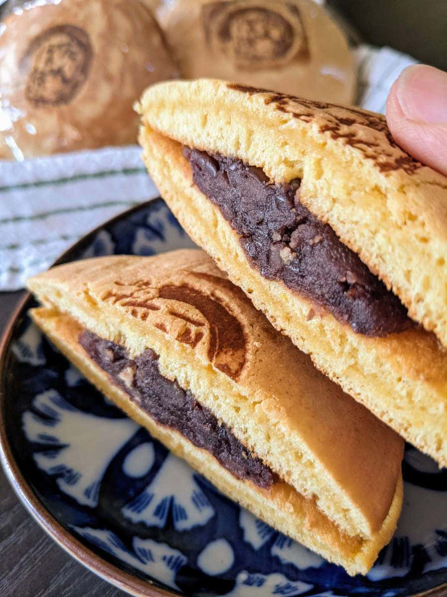 Japanese Sweets 桌布 壁紙 — 一隻手拿著半個金黃色煎餅，露出豐富的深紅色豆沙餡，放在藍白圖案盤上。