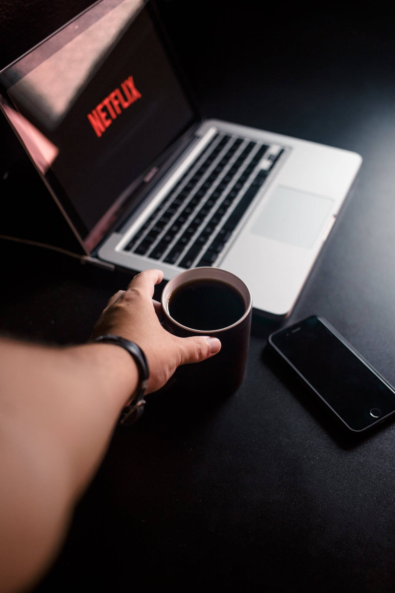 深色 iPhone 桌布 壁紙 — 一隻手拿著咖啡杯，旁邊是一部顯示Netflix的筆記型電腦，放在深色桌子上，附近有一部手機。