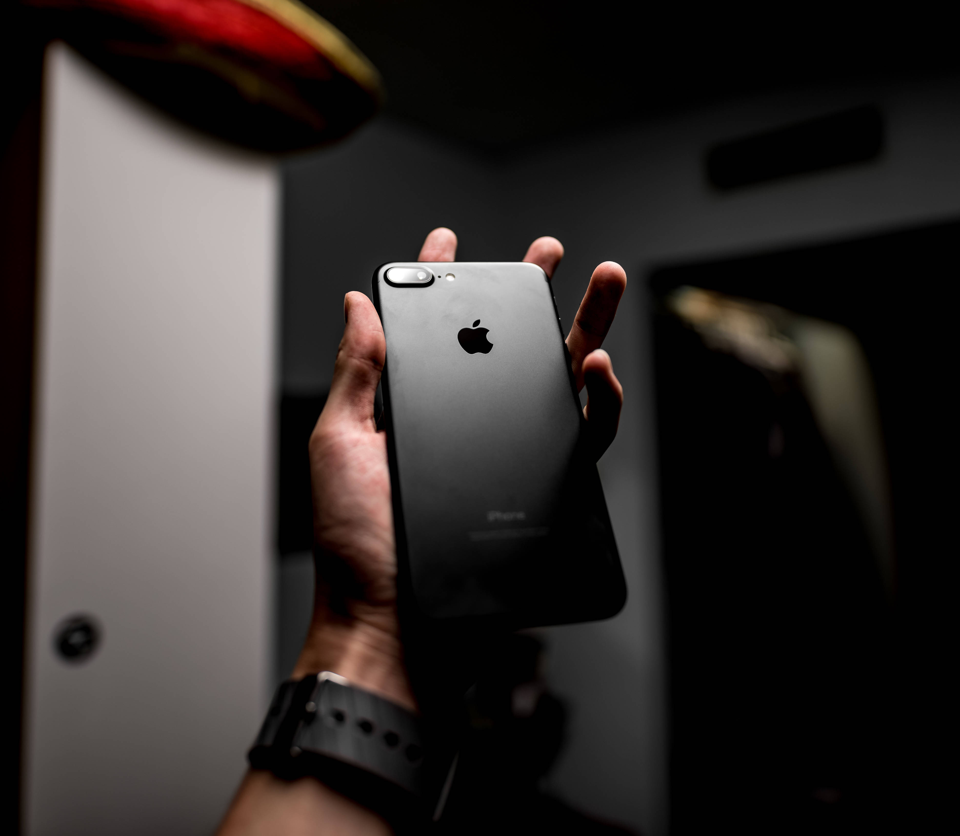 iPhone 8 桌布 壁紙 — 一隻手拿著帶有Apple標誌的黑色iPhone，背景是暗色模糊的柔和燈光。