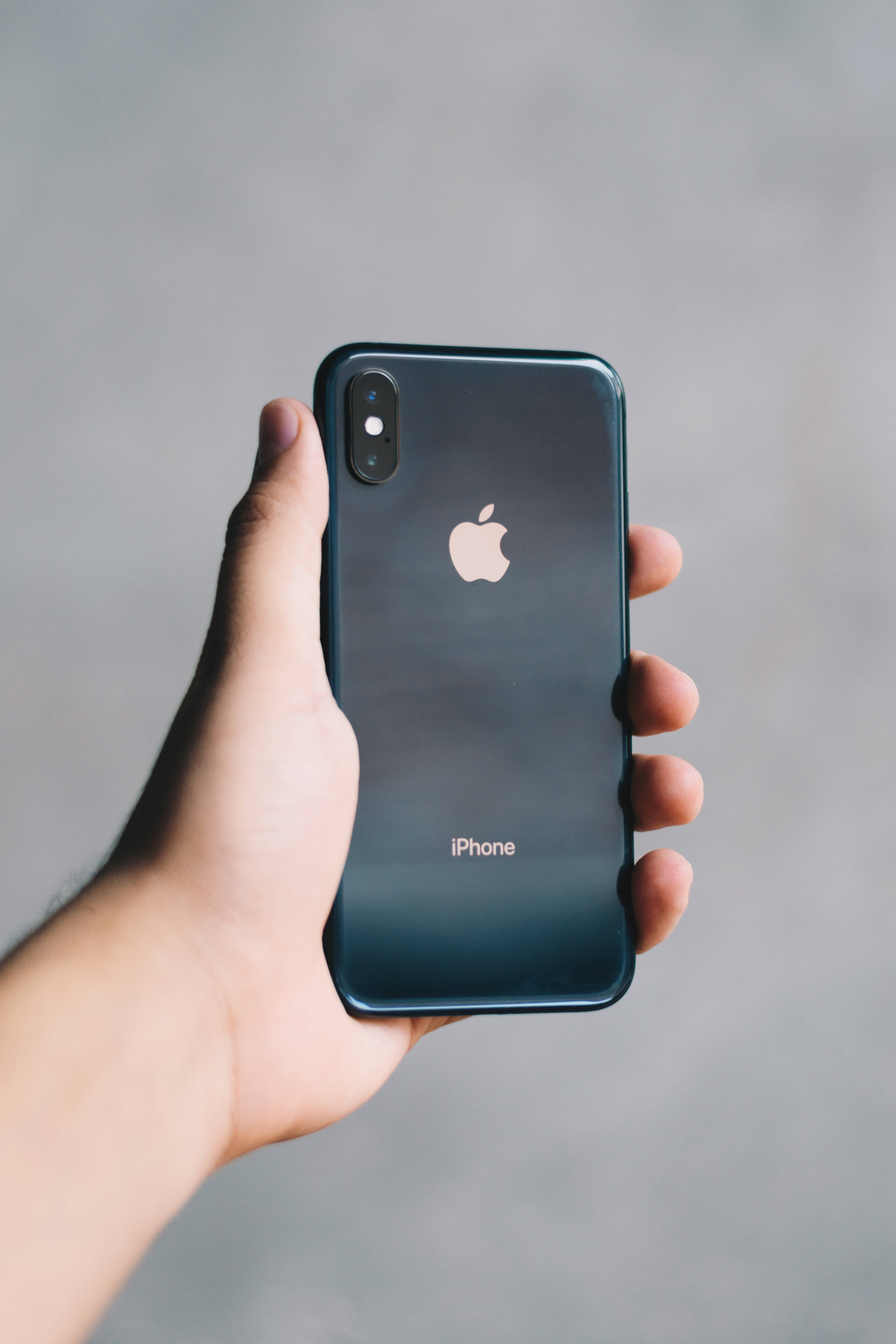 iPhone XS 桌布 壁紙 — 一隻手拿著帶有光澤表面的深藍色 iPhone X，背景為中性灰色。
