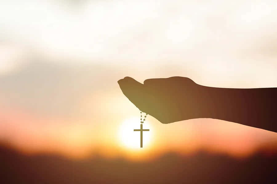 Gospel 桌布 壁紙 — 一隻手拿著帶有十字架的玫瑰經念珠，背景是閃耀的日落天空。