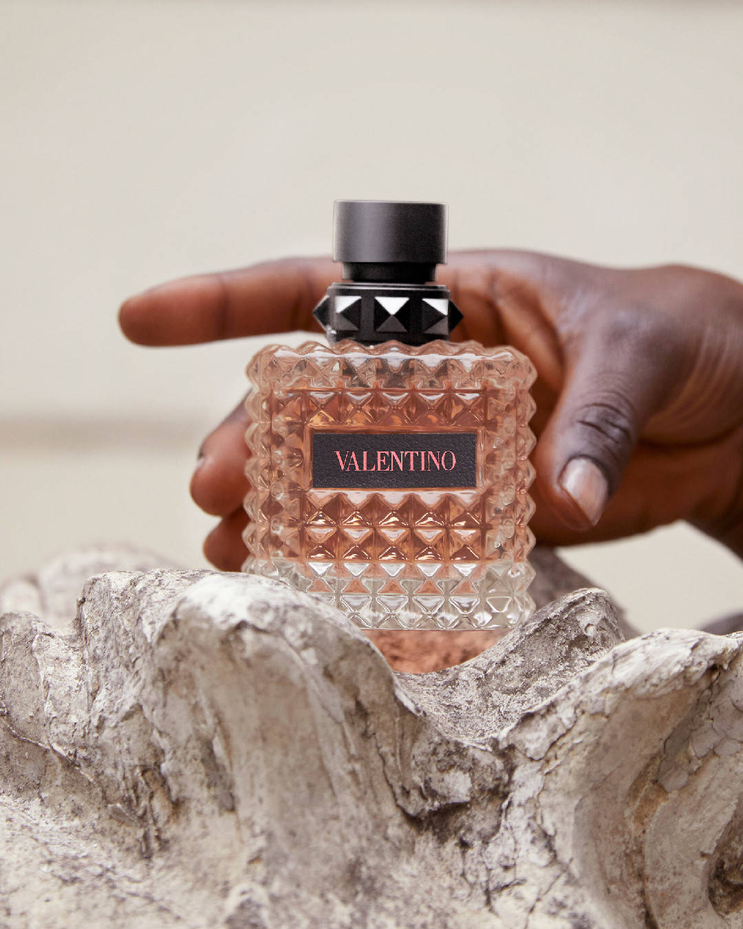 品牌 桌布 壁紙 — 一隻手拿著帶有紋理的 Valentino 香水瓶，內有玫瑰金色液體，背景為中性色調。