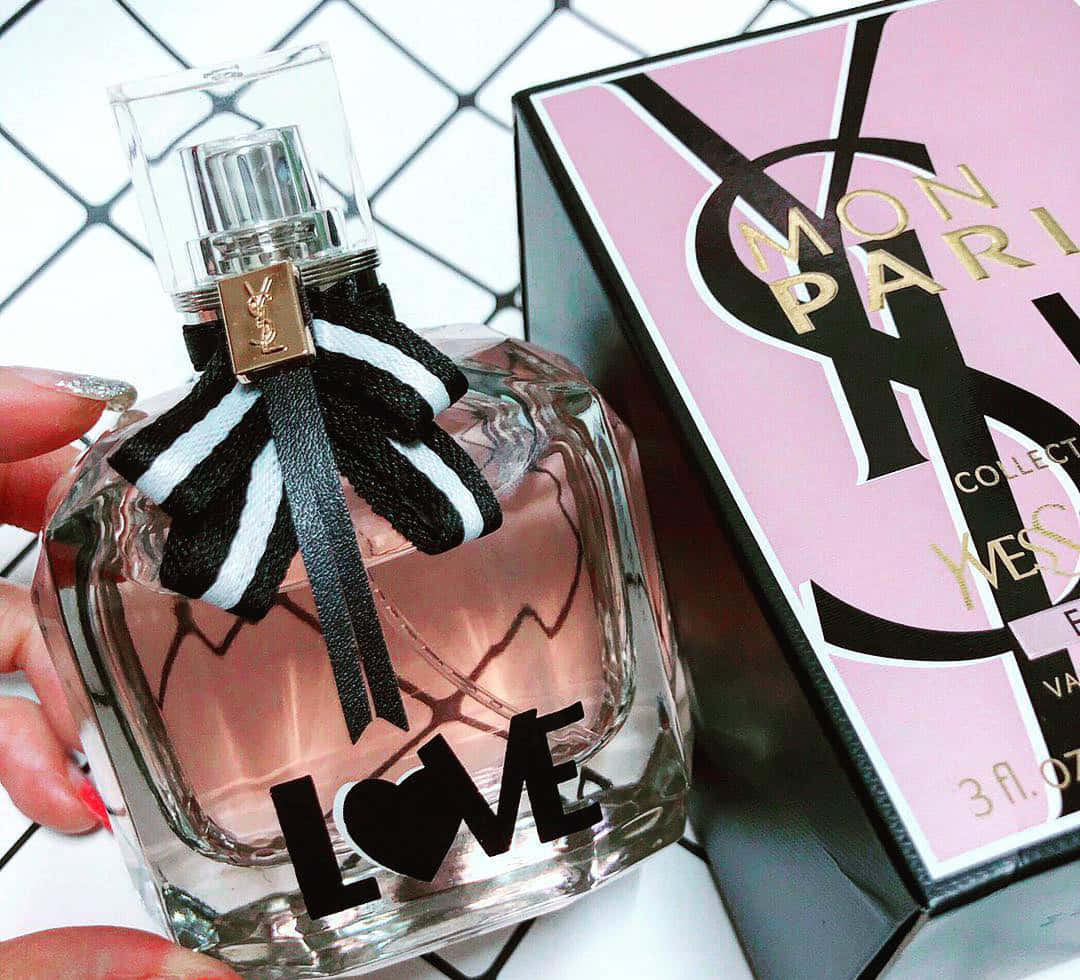 Ysl 桌布 壁紙 — 一隻手拿著帶有黑白條紋蝴蝶結的YSL Mon Paris香水瓶，背景為幾何白色。