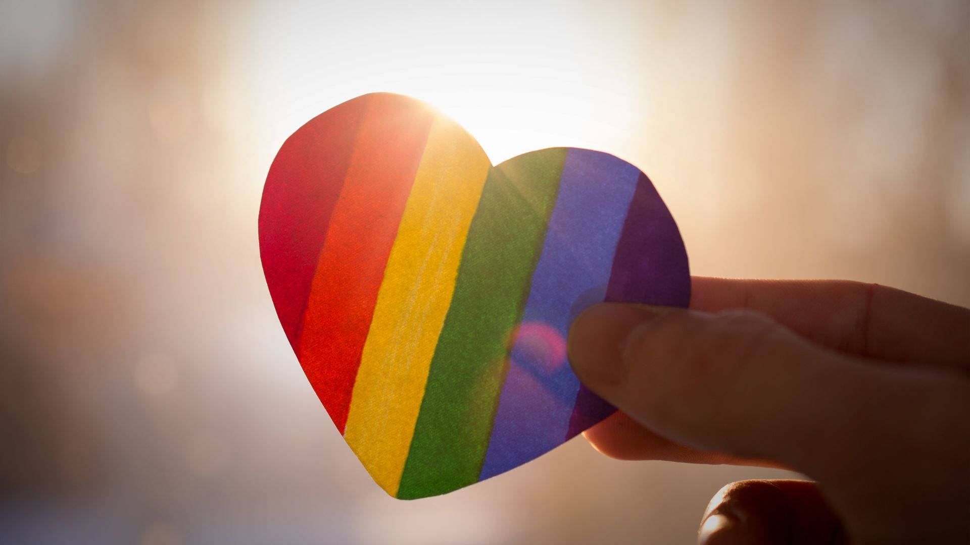 Subtle Lgbt 桌布 壁紙 — 一隻手拿著彩虹色的心形剪紙，背景是明亮的陽光和柔和散景。