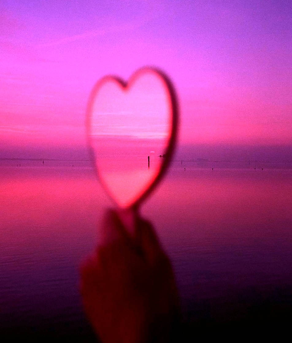Aesthetic Sunset 桌布 壁紙 — 一隻手拿著心形鏡子，映照出平靜水面上充滿活力的粉紅色和紫色日落。