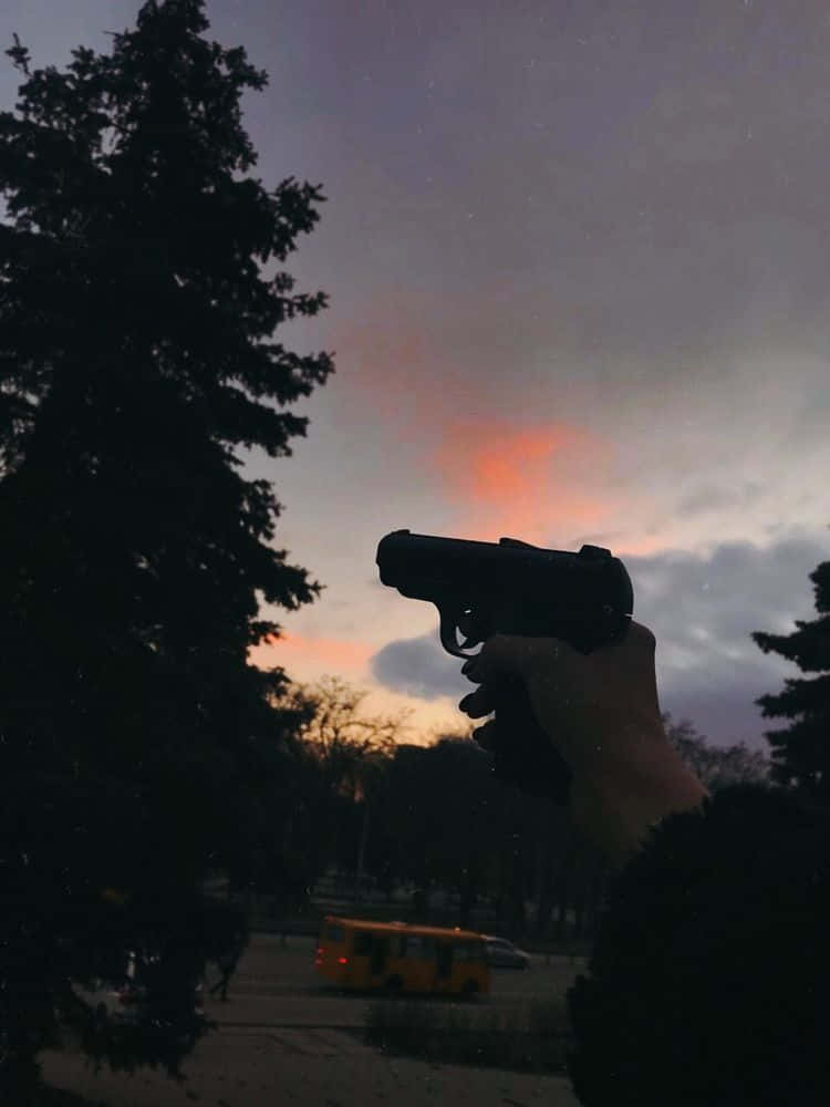 槍枝 桌布 壁紙 — 一隻手拿著手槍，背景是帶有橙色雲彩和剪影樹木的黃昏天空。