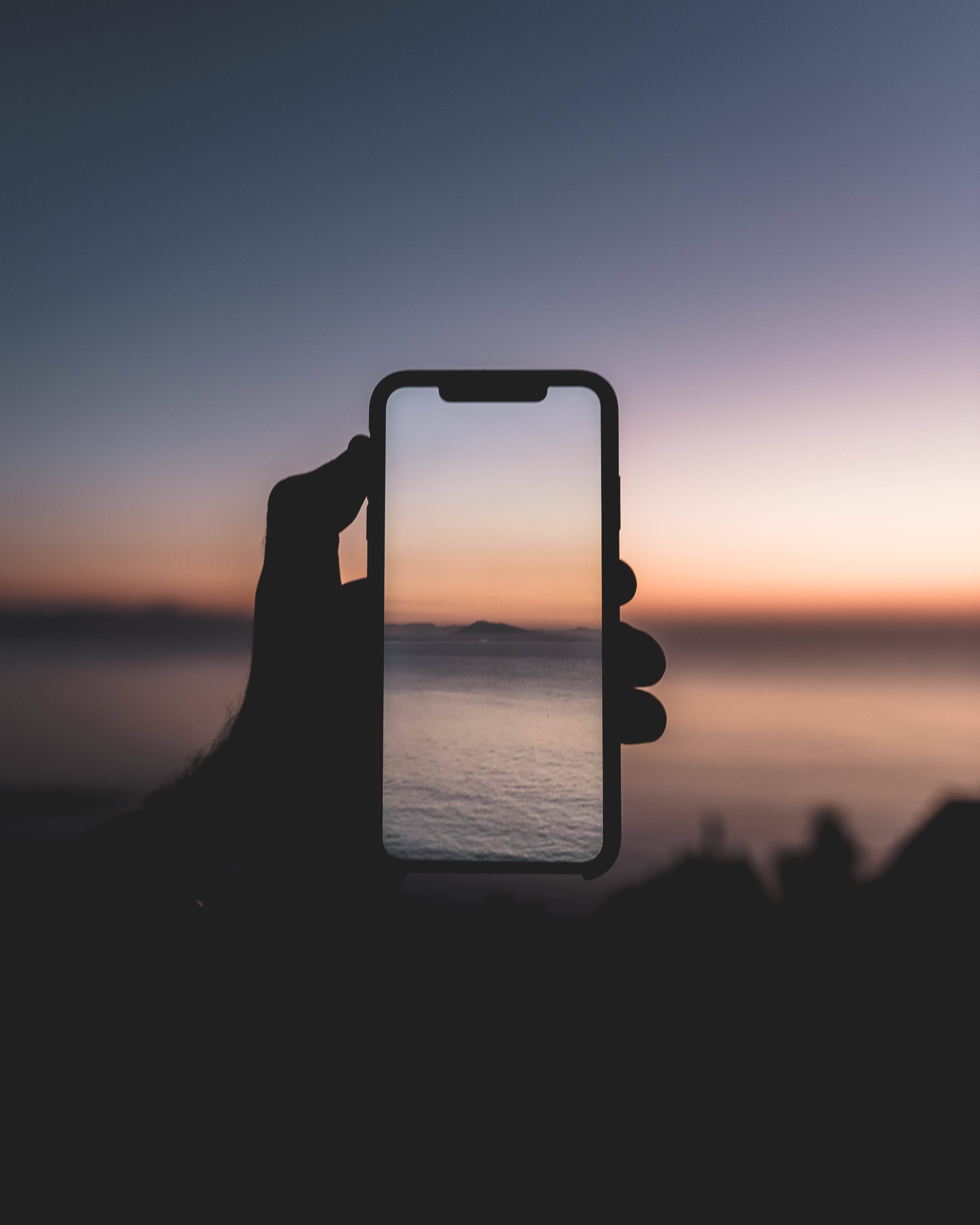 4K iPhone 11 桌布 壁紙 — 一隻手拿著手機，捕捉水面上一片寧靜的日落，帶有柔和的橙色和紫色色調。