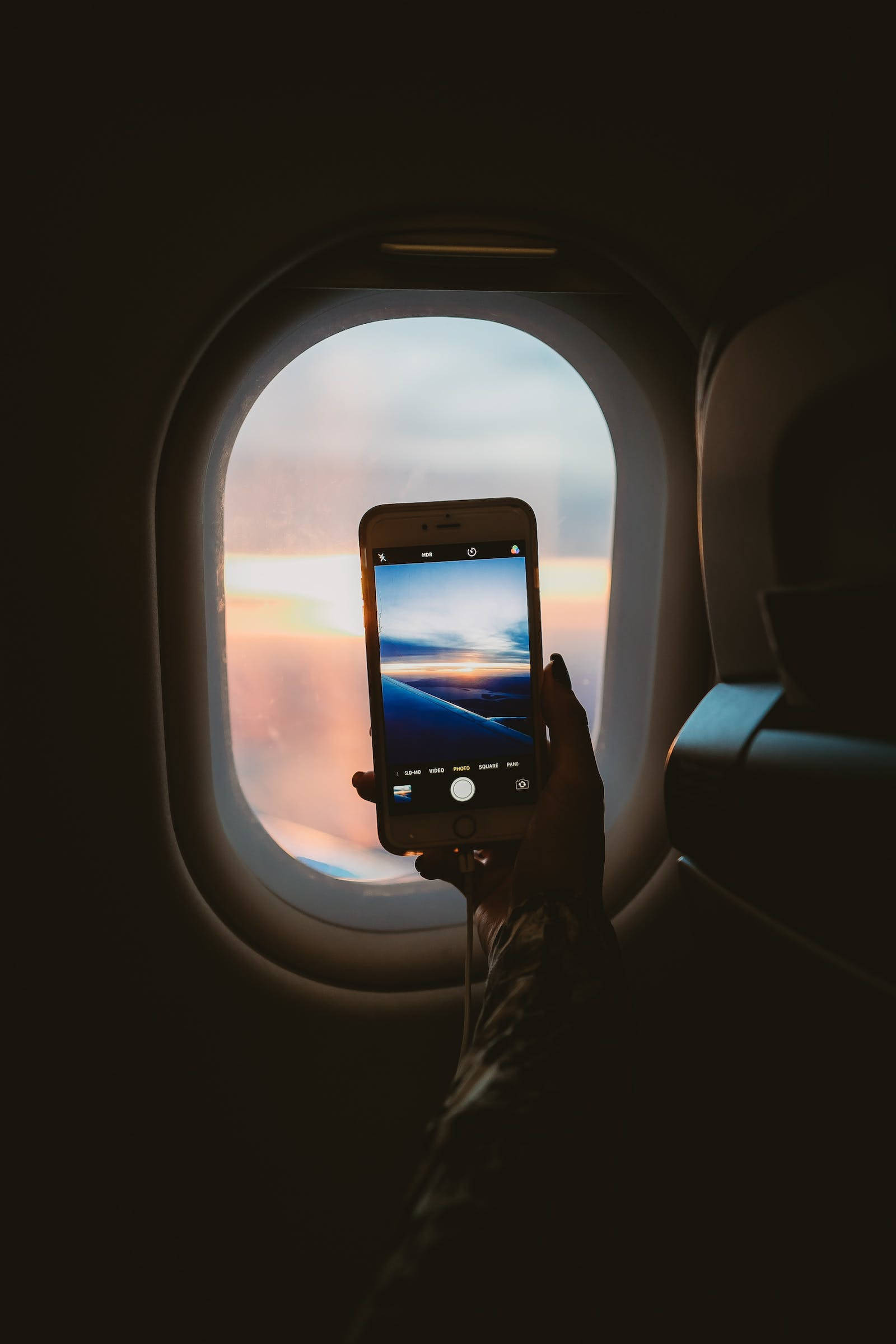 深色 iPhone 桌布 壁紙 — 一隻手拿著手機，透過飛機窗戶捕捉充滿活力的日落，伴隨著柔軟的雲朵和溫暖的色調。
