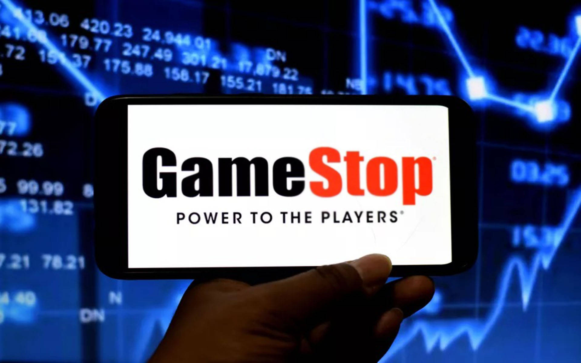 GameStop 桌布 壁紙 — 一隻手拿著手機，顯示GameStop標誌，背景是發光的藍色股市數據。