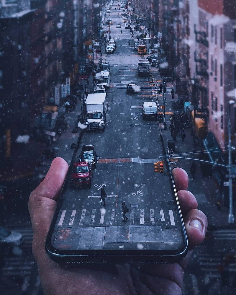 Digital 桌布 壁紙 — 一隻手拿著手機，顯示一條積雪的街道，有汽車和行人在飄落的雪花下。