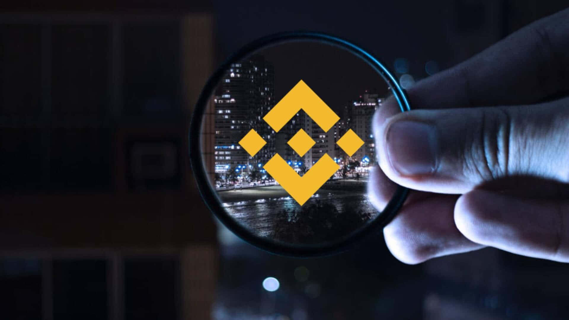 Binance 桌布 壁紙 — 一隻手拿著放大鏡在夜晚的城市景觀上，露出一個發光的黃色Binance標誌。