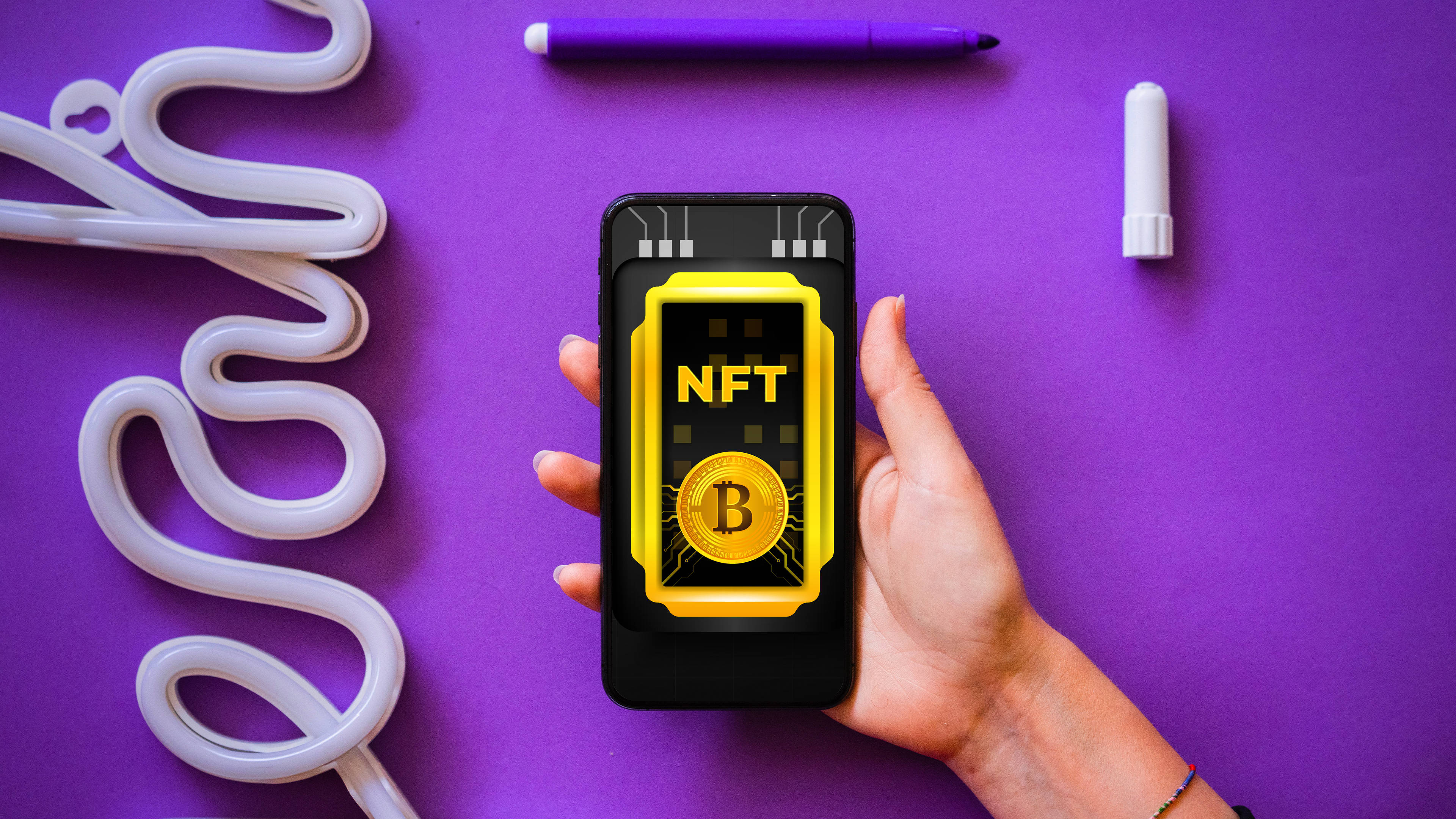 NFT 桌布 壁紙 — 一隻手拿著智慧型手機，在充滿活力的紫色背景上顯示金色NFT代幣，並有白色霓虹管和筆。