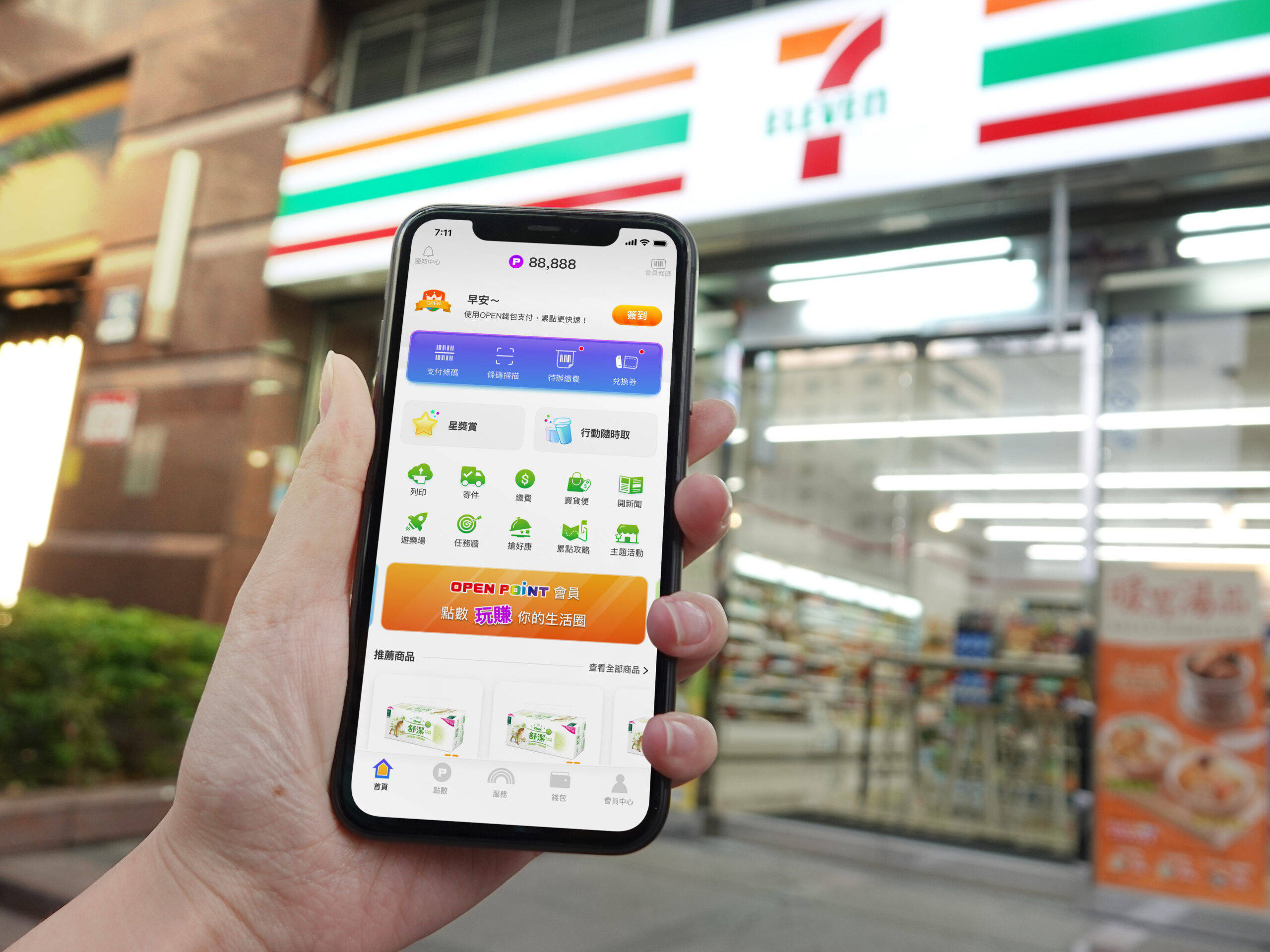 Eleven 桌布 壁紙 — 一隻手拿著智慧型手機，在夜晚燈光明亮的便利商店前顯示7-Eleven應用程式介面。