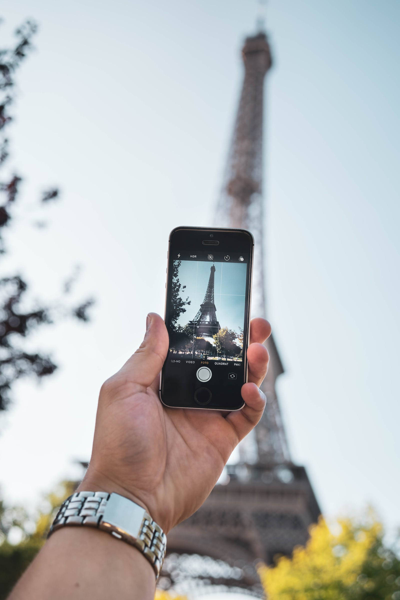 france iPhone 桌布 壁紙 — 一隻手拿著智慧型手機，在明亮的天空和模糊的秋樹背景下拍攝艾菲爾鐵塔。