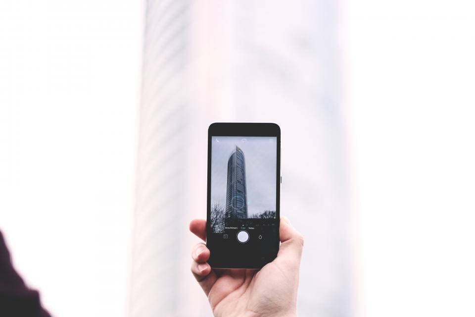 photography iPhone 桌布 壁紙 — 一隻手拿著智慧型手機，在明亮的陰天背景下拍攝一座高聳的現代摩天大樓。