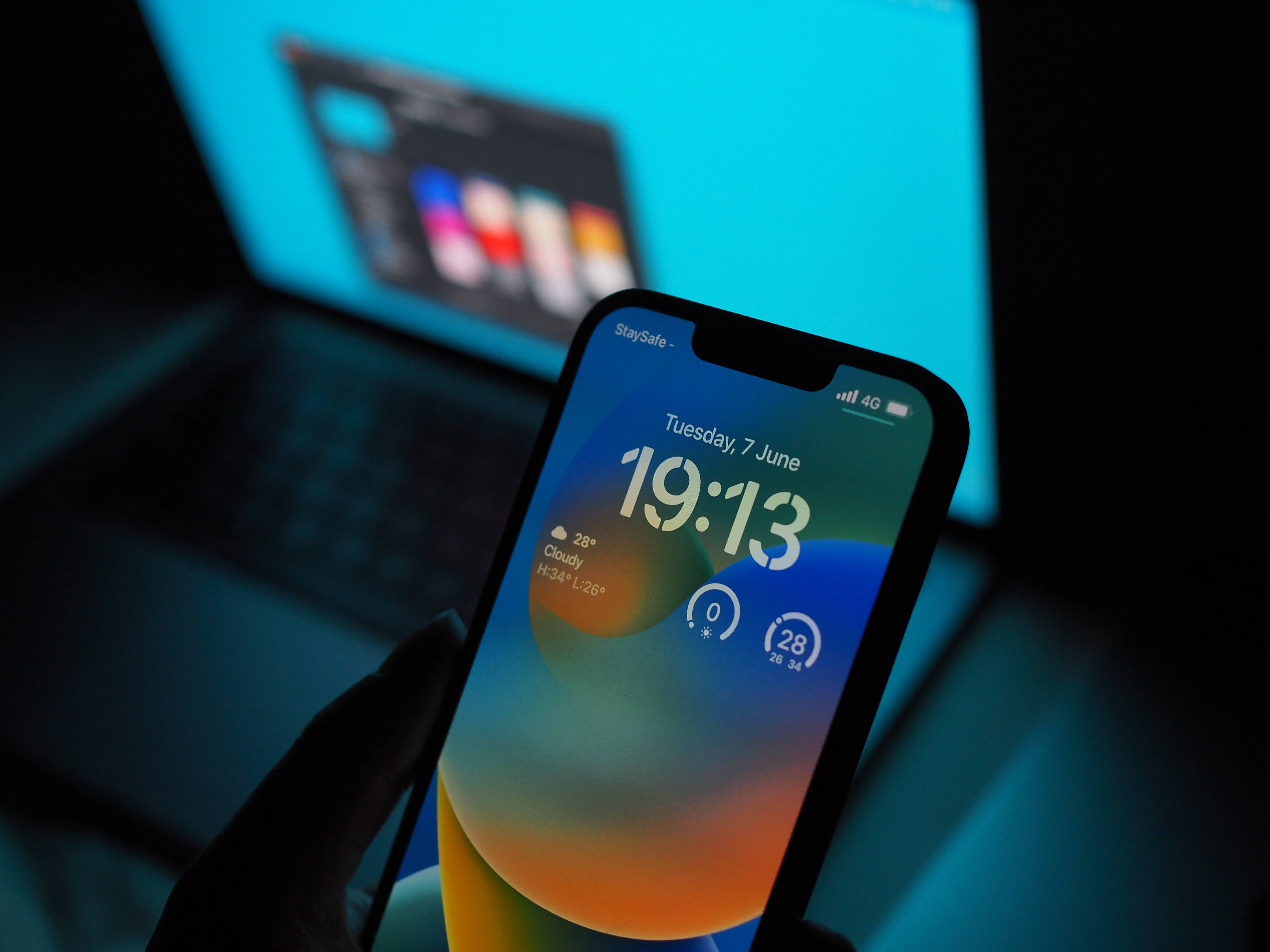 iOS 16 壁紙 桌布 壁紙 — 一隻手拿著智慧型手機，在昏暗的筆記型電腦背景上顯示充滿活力的漸變桌布，包含時間、天氣和日期。