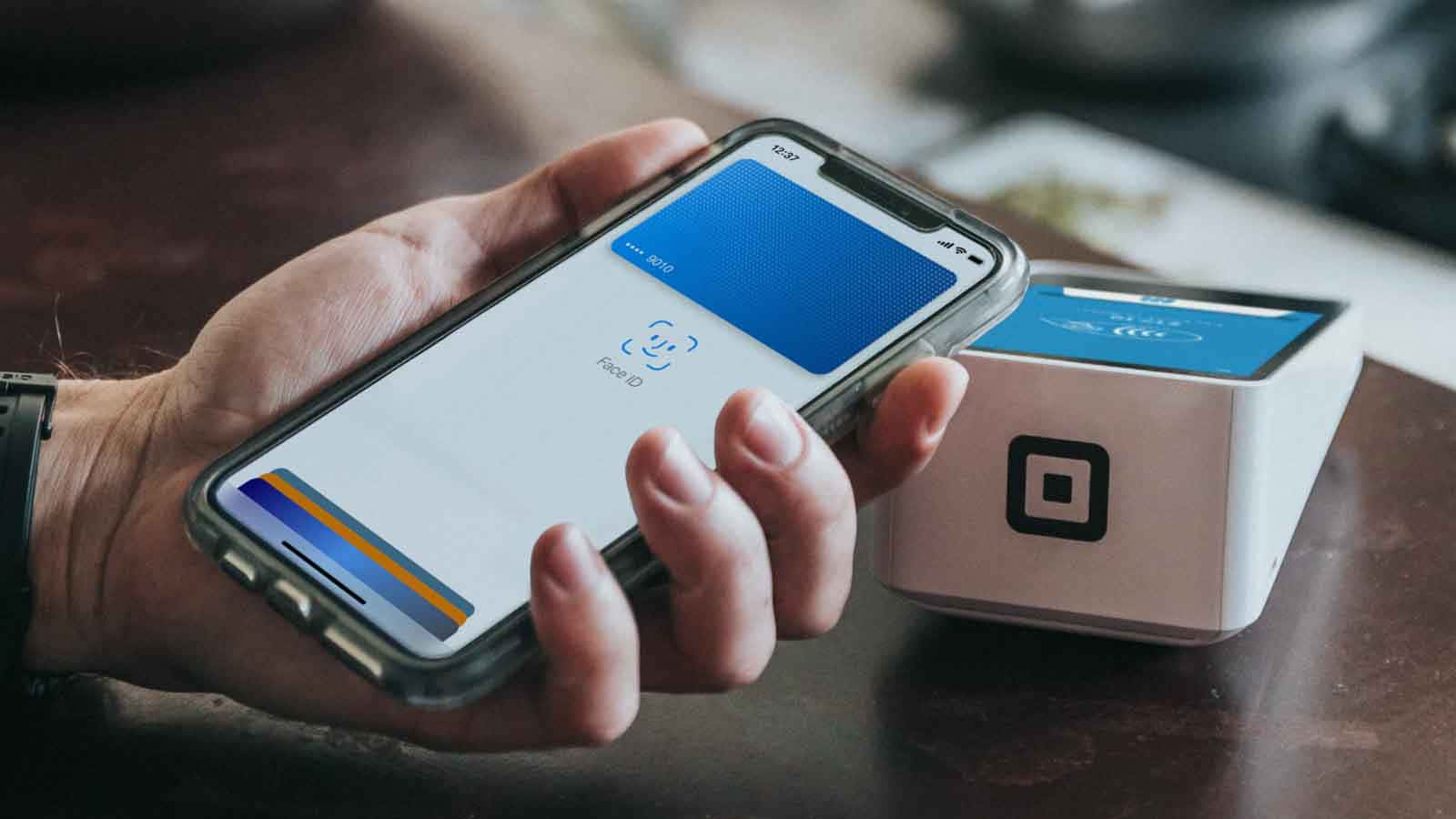 Smartphone 桌布 壁紙 — 一隻手拿著智慧型手機，在木桌上靠近Square讀卡機顯示非接觸式付款畫面。