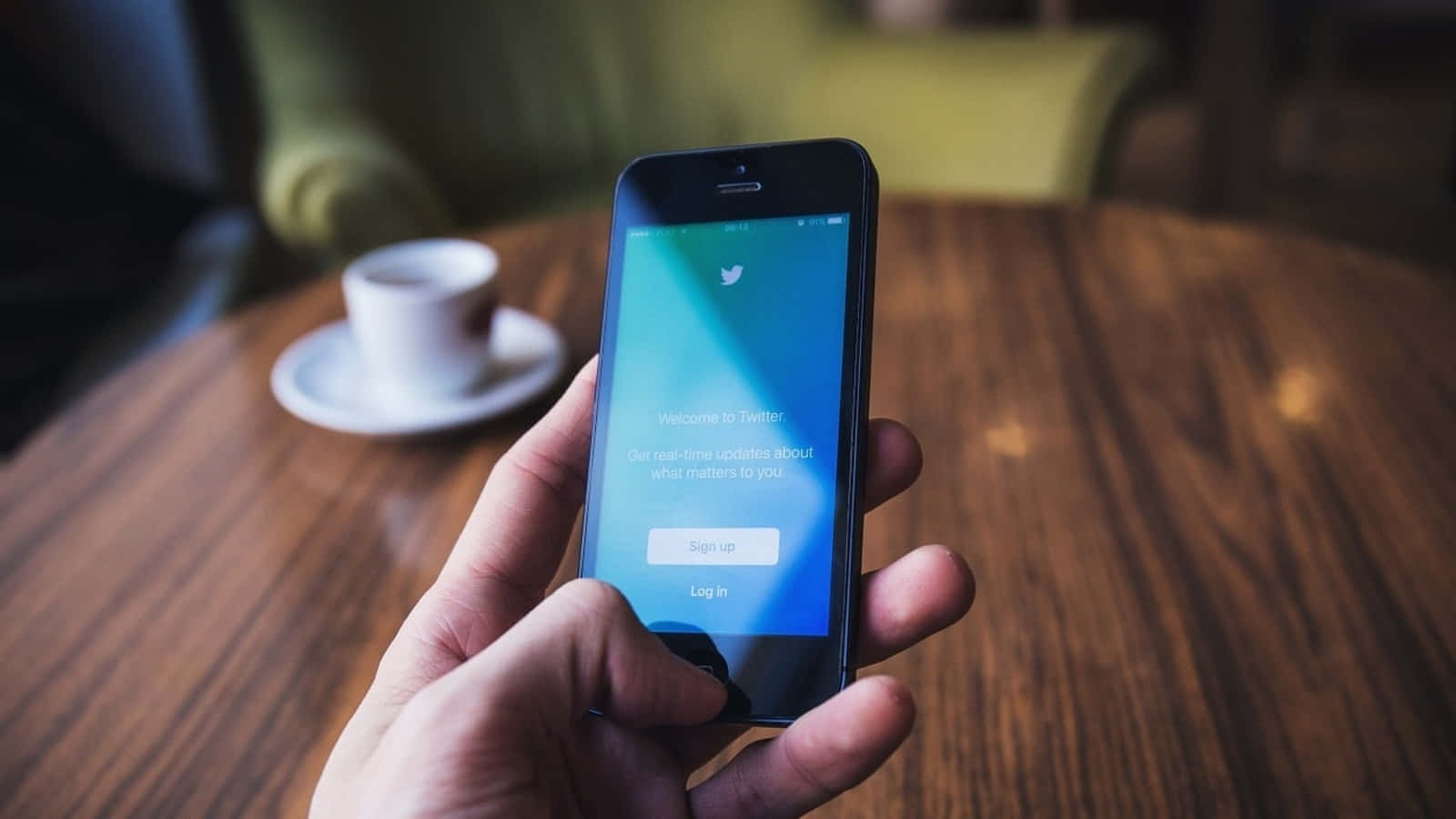 Twitter 桌布 壁紙 — 一隻手拿著智慧型手機，在木桌上顯示Twitter的歡迎畫面，背景有柔焦的咖啡杯。