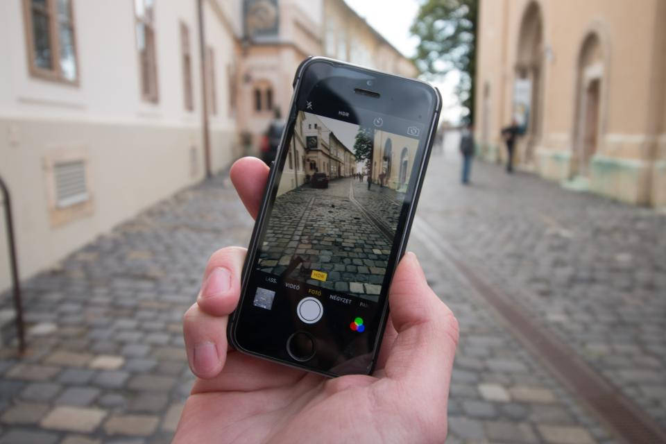 photography iPhone 桌布 壁紙 — 一隻手拿著智慧型手機，在柔和日光下拍攝一條兩旁歷史建築的鵝卵石街道。