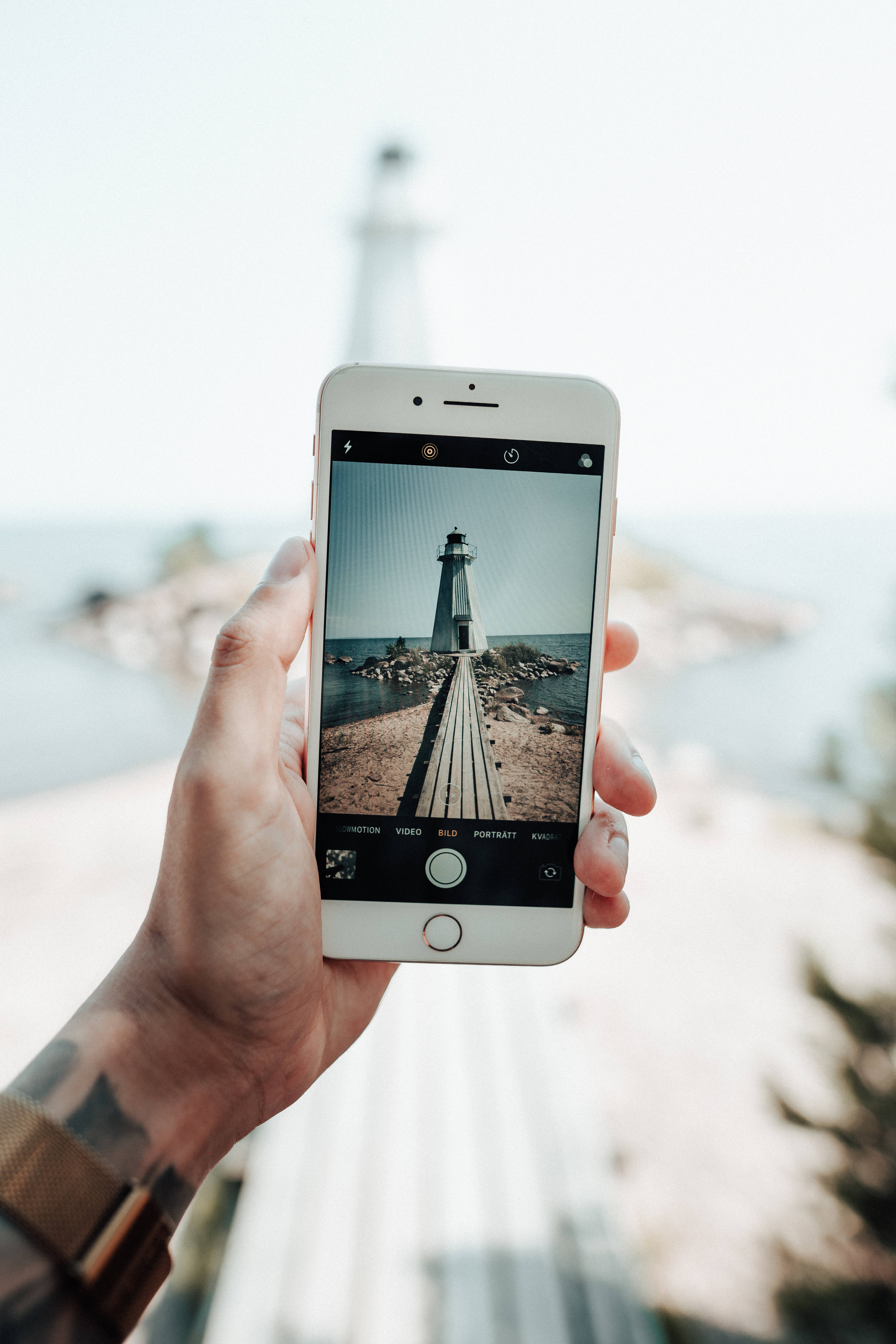 photography iPhone 桌布 壁紙 — 一隻手拿著智慧型手機，在柔和日光下拍攝平靜海面上木碼頭盡頭的燈塔。