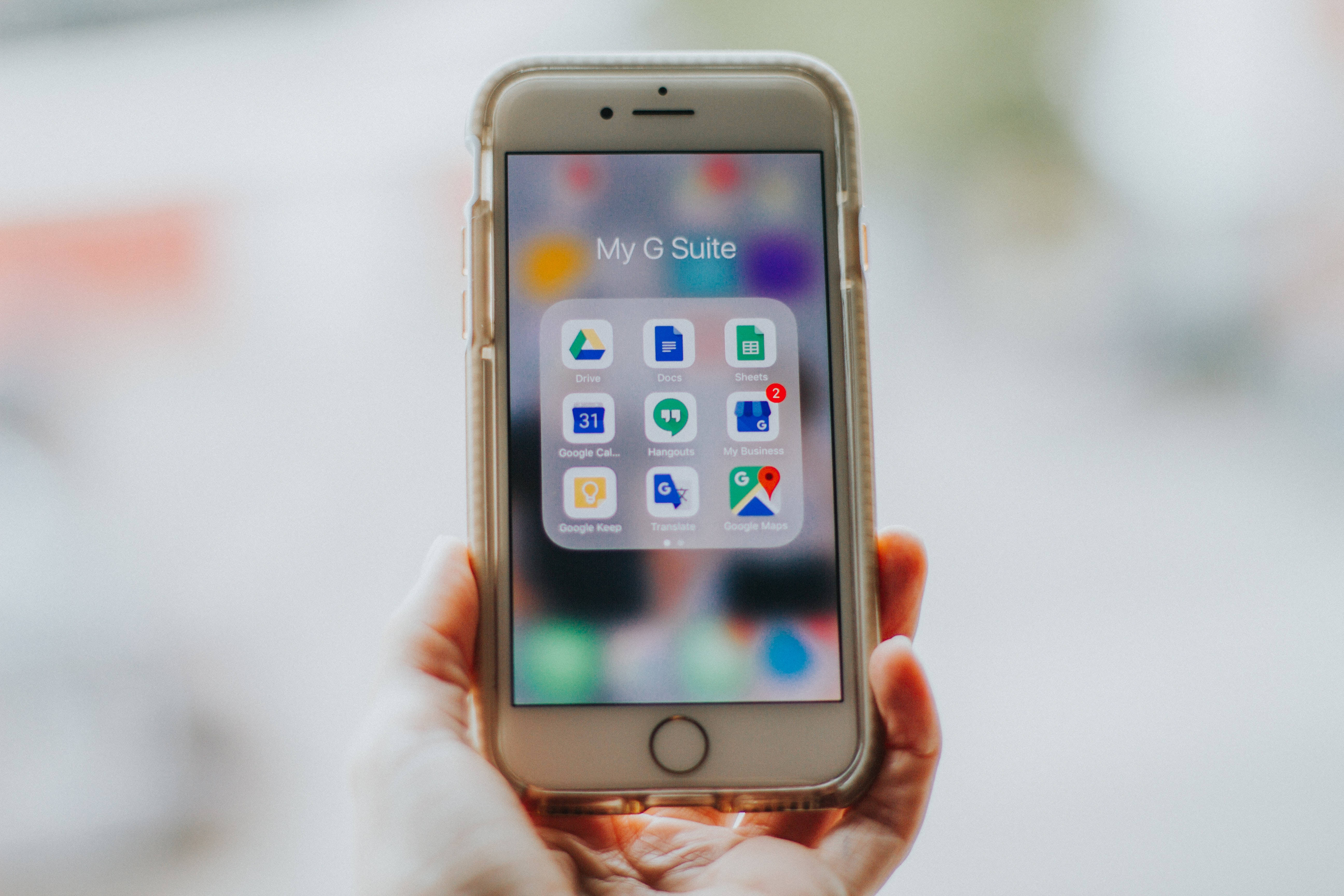 Software 桌布 壁紙 — 一隻手拿著智慧型手機，在模糊的背景上顯示帶有彩色圖標的 Google G Suite 應用程式網格。