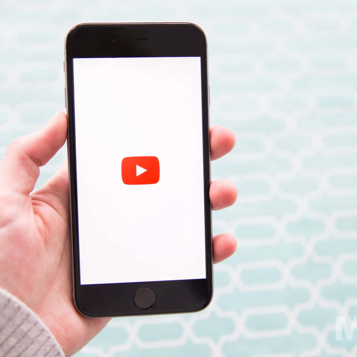 YouTube 桌布 壁紙 — 一隻手拿著智慧型手機，在淺藍色圖案背景上顯示YouTube應用程式圖示。