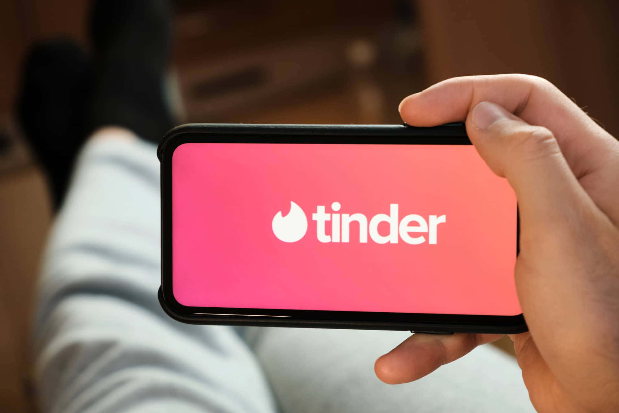 tinder 桌布 壁紙 — 一隻手拿著智慧型手機，在粉紅色漸層背景上顯示Tinder應用程式標誌。