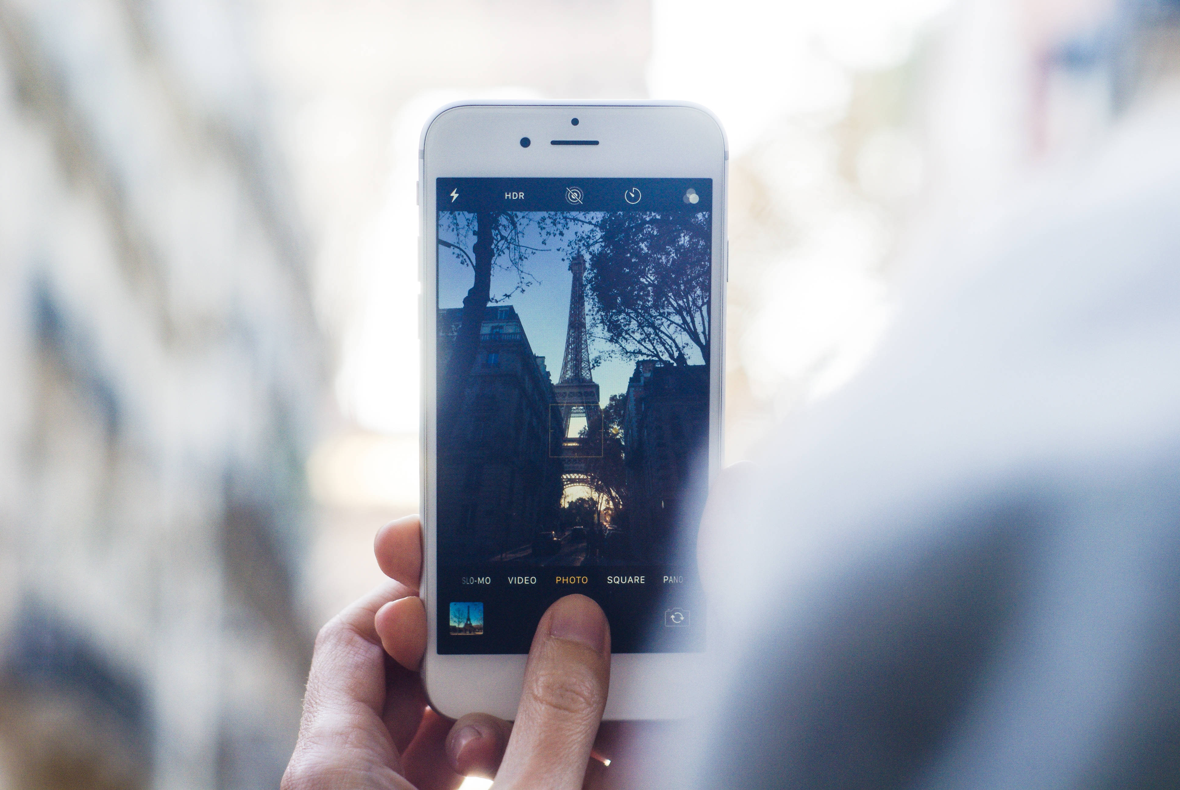 france iPhone 桌布 壁紙 — 一隻手拿著智慧型手機，在黃昏時分透過綠樹成蔭的街道，在溫暖的環境光線下拍攝艾菲爾鐵塔。