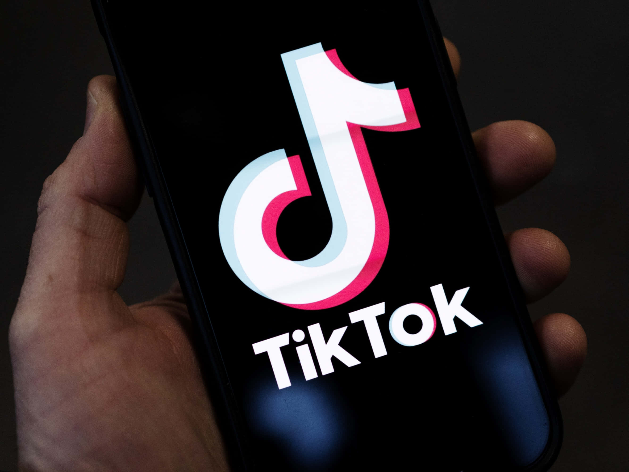 TikTok 動圖 桌布 壁紙 — 一隻手拿著智慧型手機，在黑暗背景上顯示明亮的TikTok標誌。