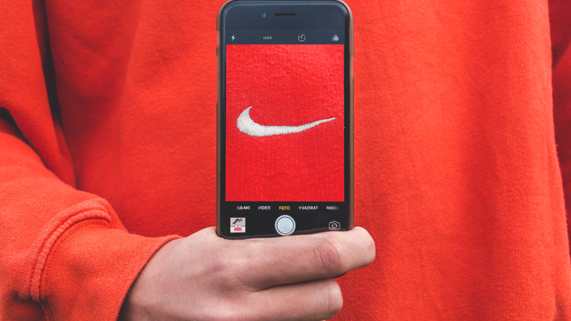 Nike iPhone 桌布 壁紙 — 一隻手拿著智慧型手機，拍攝在鮮紅色布料上刺繡的白色Nike標誌的特寫。