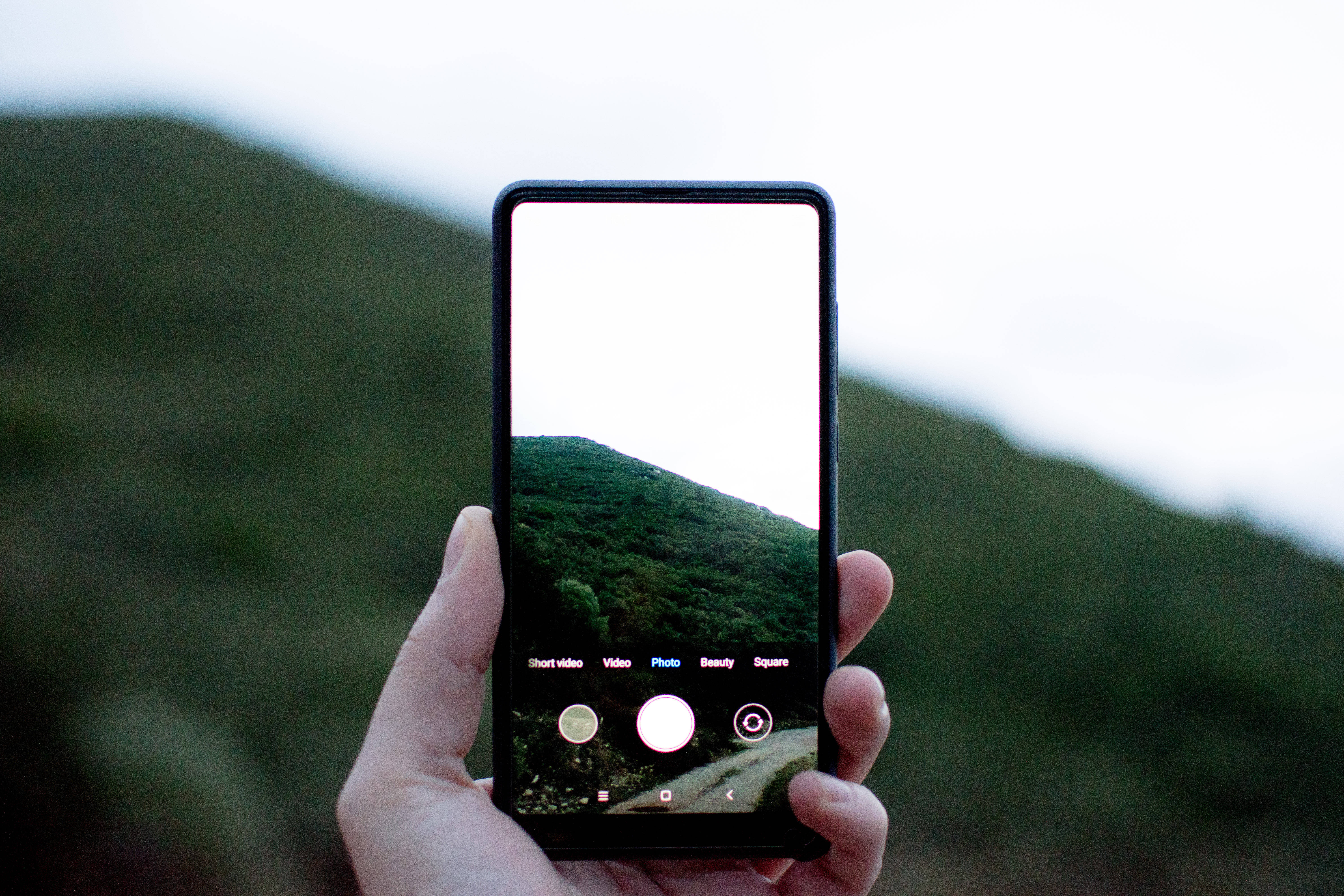 Xiaomi 桌布 壁紙 — 一隻手拿著智慧型手機，拍攝陰天下一片翠綠的山丘。