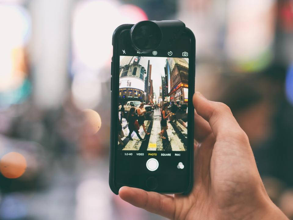 photography iPhone 桌布 壁紙 — 一隻手拿著智慧型手機，捕捉繁忙的城市街道場景，行人正在明亮的燈光下穿越斑馬線。