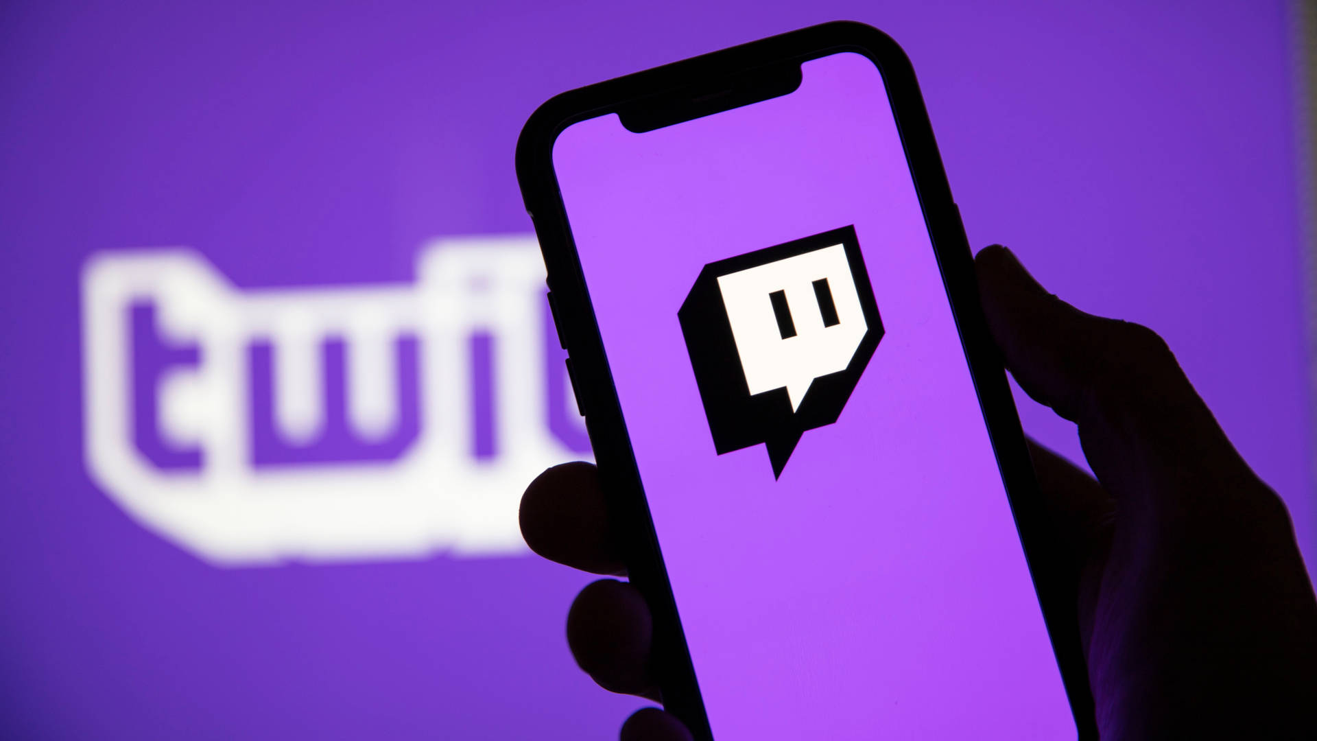 Twitch 1080 桌布 壁紙 — 一隻手拿著智慧型手機，螢幕上顯示 Twitch 標誌，背景為紫色，後面有模糊的 Twitch 標誌。