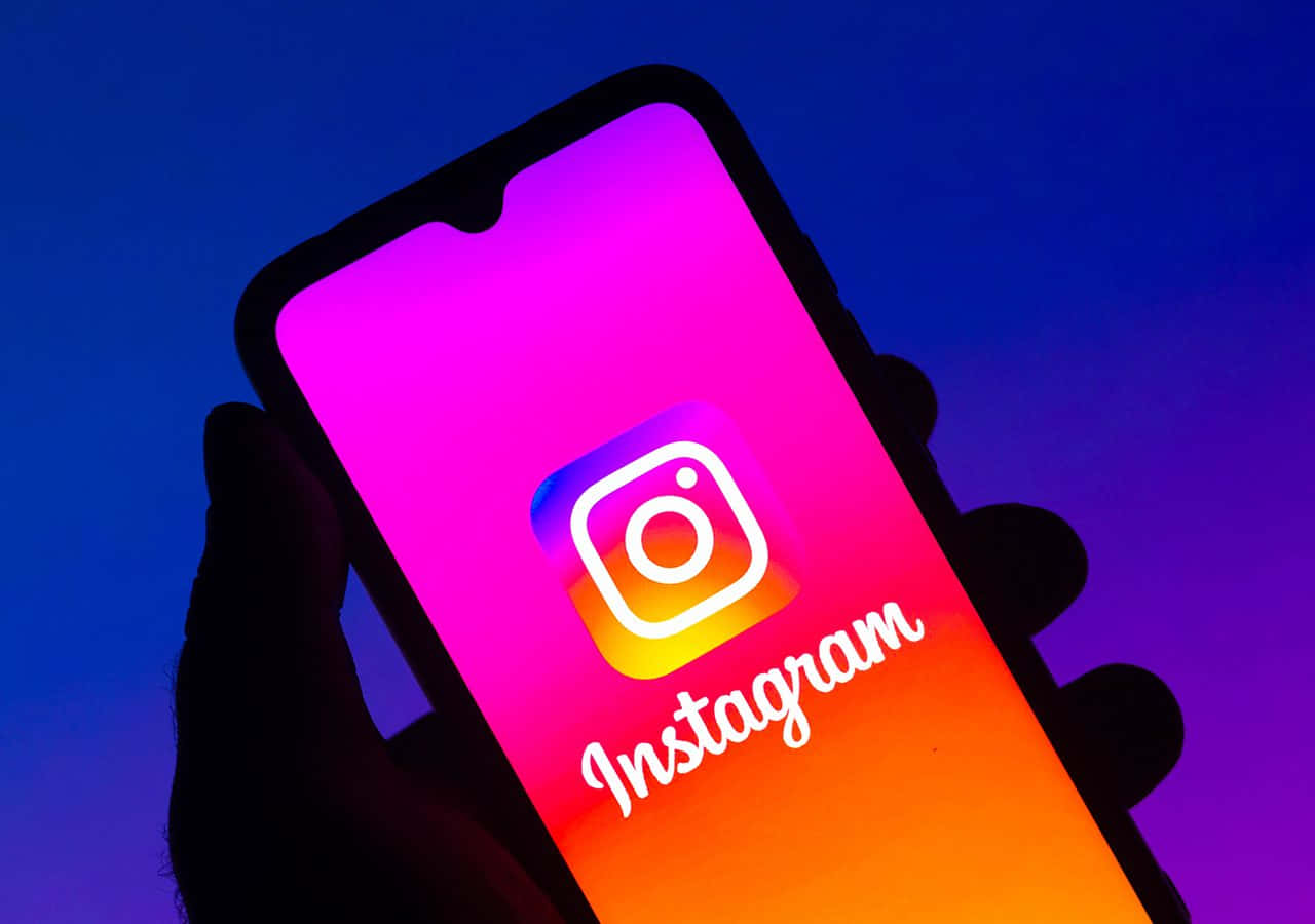 Instagram 桌布 壁紙 — 一隻手拿著智慧型手機，螢幕上顯示Instagram應用程式標誌，背景是鮮豔的紫色和橙色漸變色。