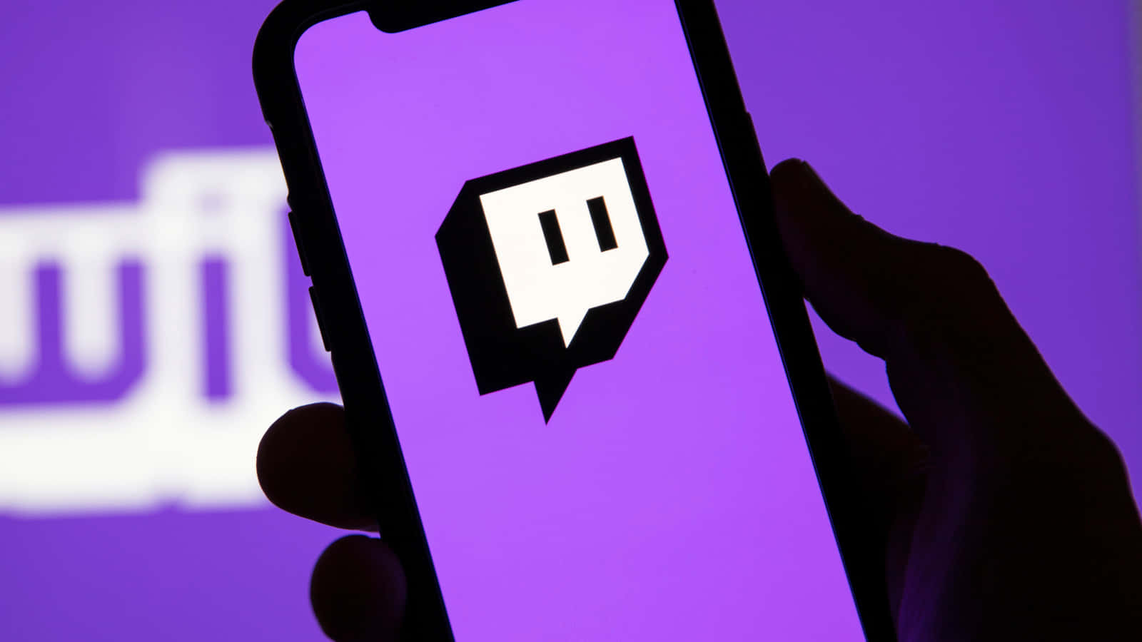 Twitch 桌布 壁紙 — 一隻手拿著智慧型手機，螢幕上顯示 Twitch 標誌，背景是鮮豔的紫色，後面有模糊的 Twitch 標誌。