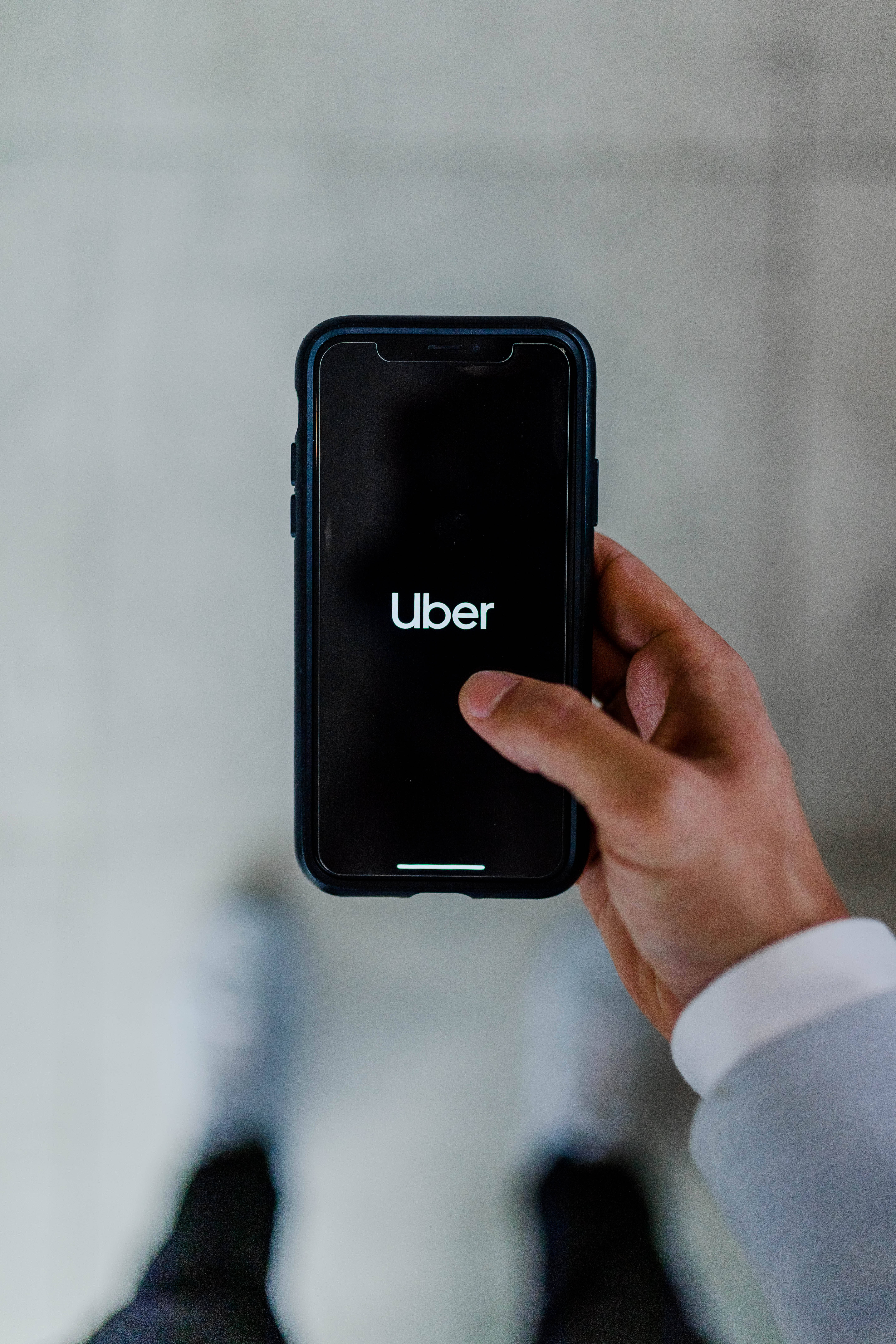 transportation 桌布 壁紙 — 一隻手拿著智慧型手機，螢幕上顯示Uber應用程式標誌，背景為模糊的混凝土。