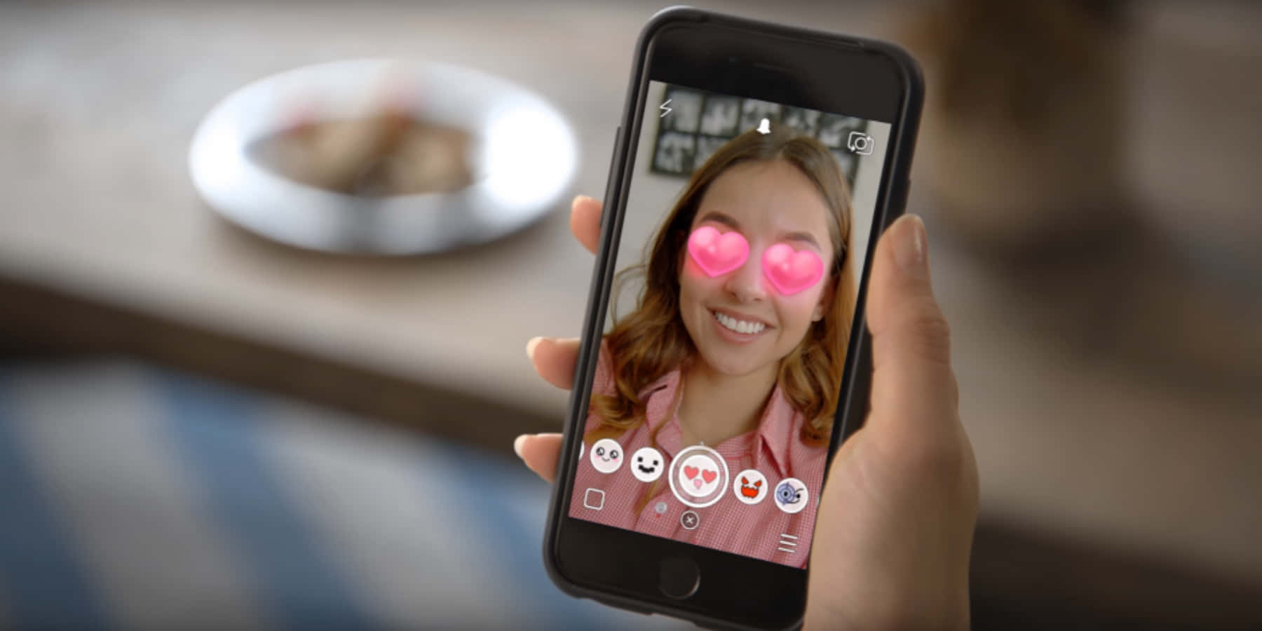 Snapchat 桌布 壁紙 — 一隻手拿著智慧型手機，螢幕上顯示一位微笑的女性，眼睛上有粉紅色心形濾鏡，身後桌上放著一個模糊的盤子。