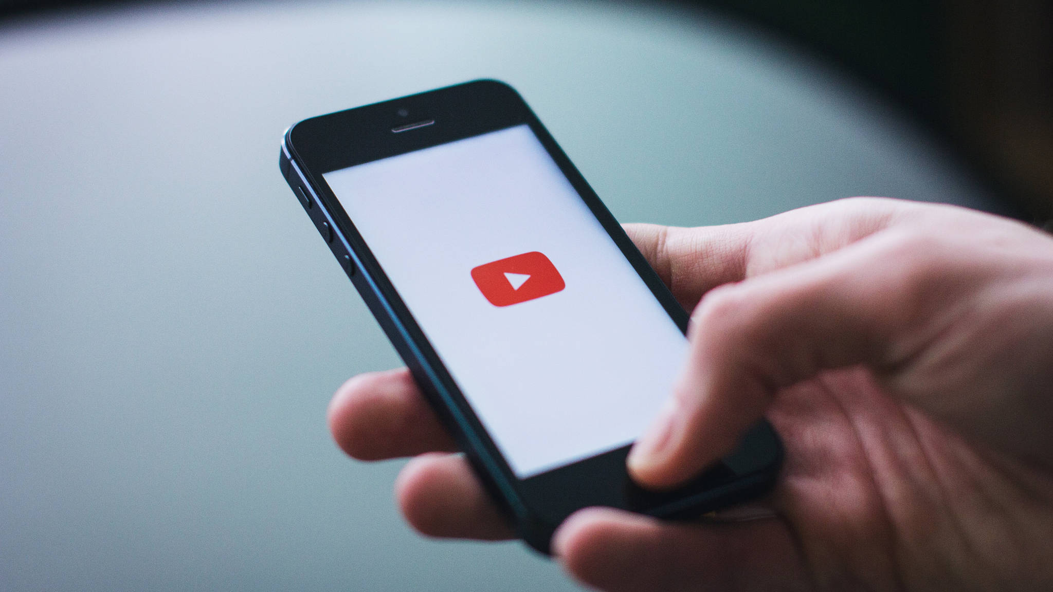 Internet 桌布 壁紙 — 一隻手拿著智慧型手機，螢幕上顯示YouTube應用程式圖示，背景為模糊的灰色。