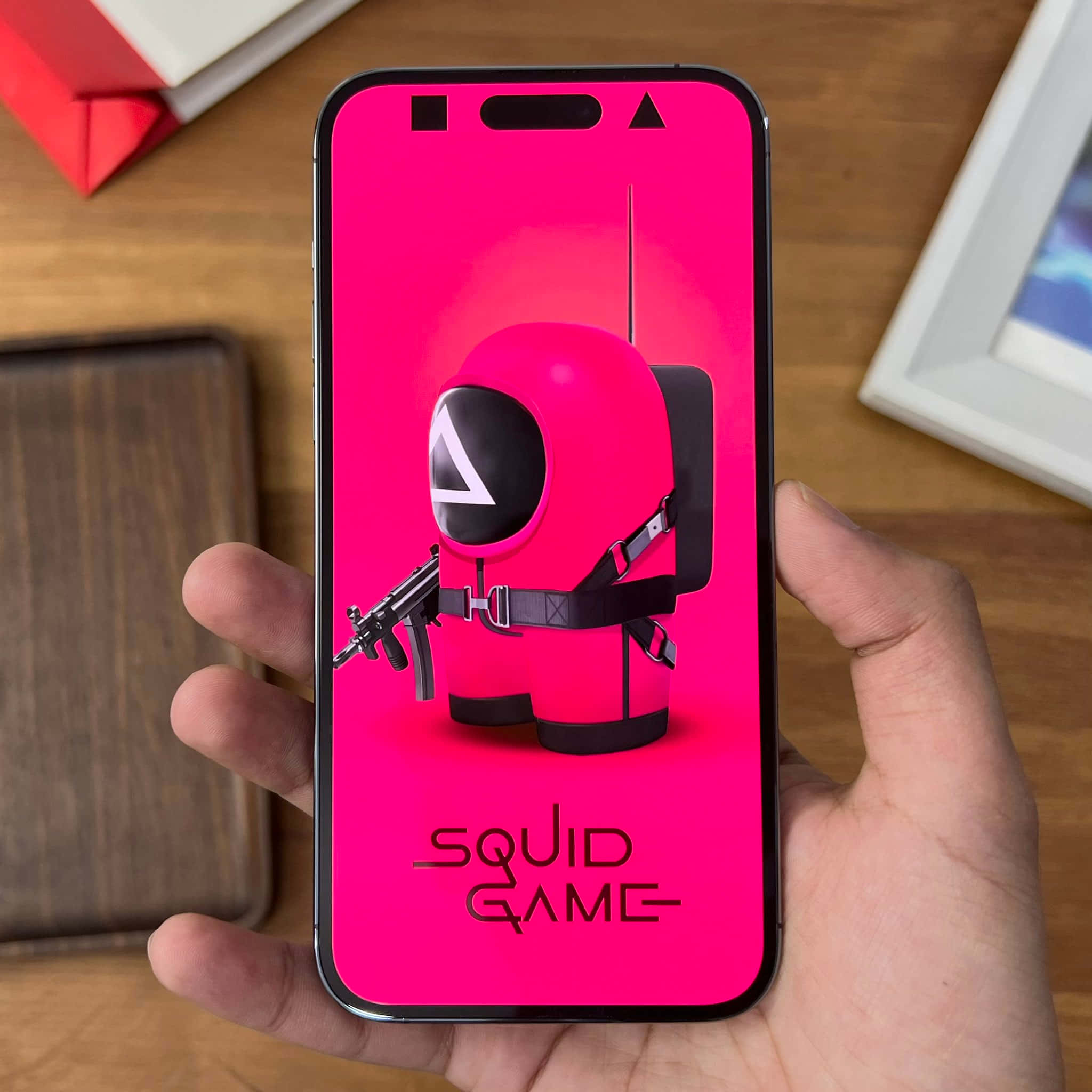 Squid Game 桌布 壁紙 — 一隻手拿著智慧型手機，螢幕上顯示一個亮粉紅色的魷魚遊戲角色，在鮮豔的粉紅色背景上持槍。