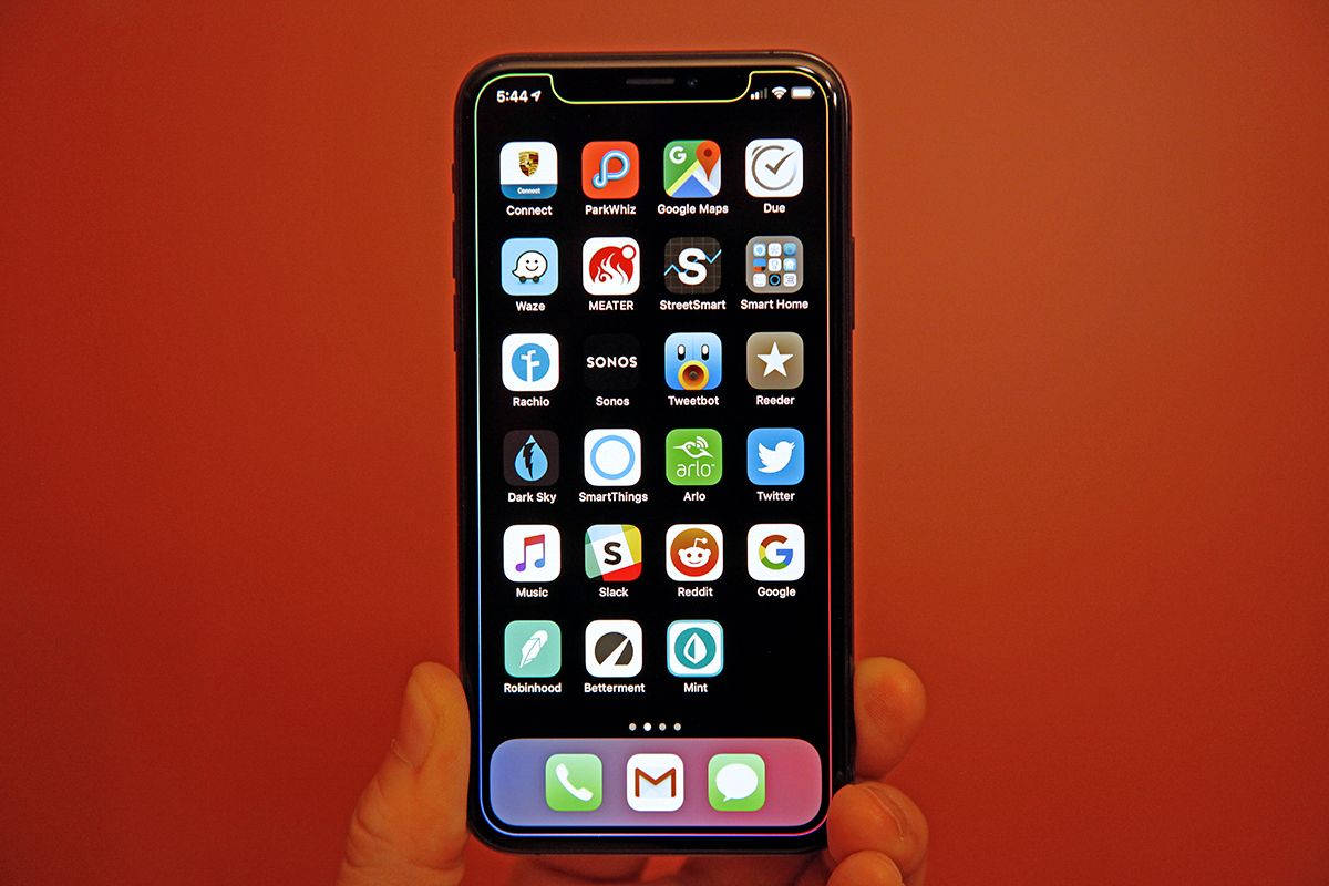 iPhone 桌布 壁紙 — 一隻手拿著智慧型手機，螢幕上顯示色彩繽紛的應用程式圖示網格，背景是溫暖的橙色牆壁。