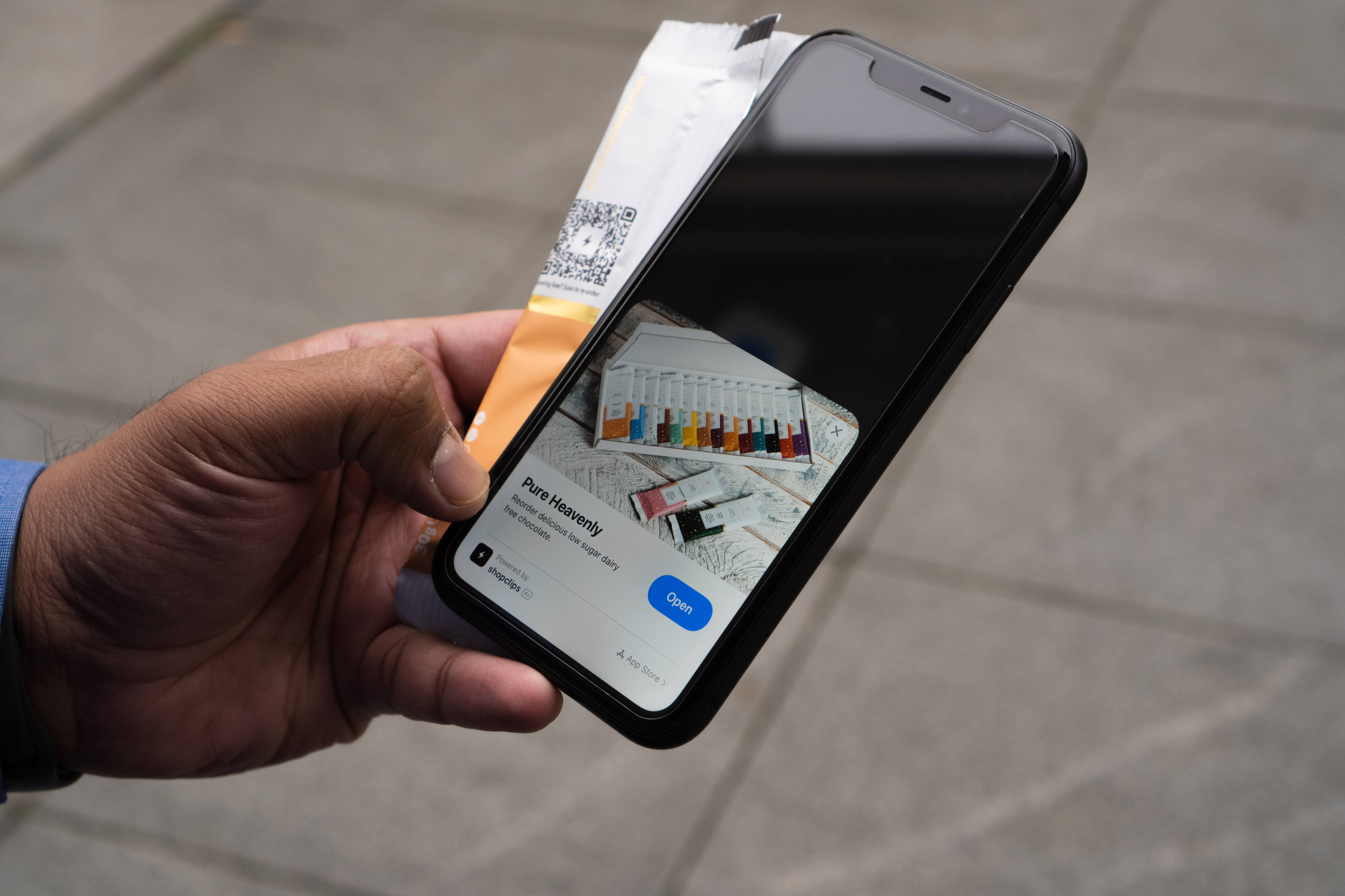 iOS 桌布 壁紙 — 一隻手拿著智慧型手機，螢幕上顯示Pure Heavenly應用程式，帶有色彩繽紛的顏料調色板和畫筆，背景是模糊的人行道。