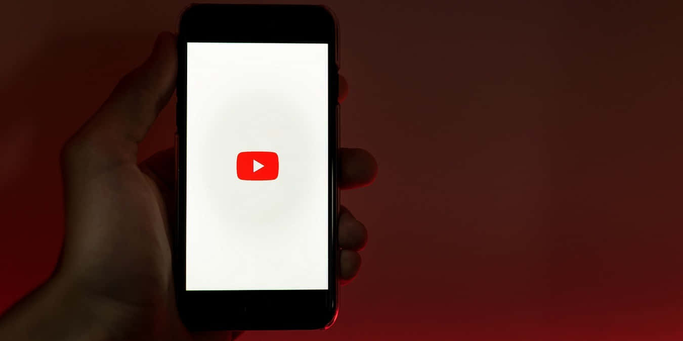 YouTube 桌布 壁紙 — 一隻手拿著智慧型手機，螢幕上顯示YouTube標誌，背景為深紅色。