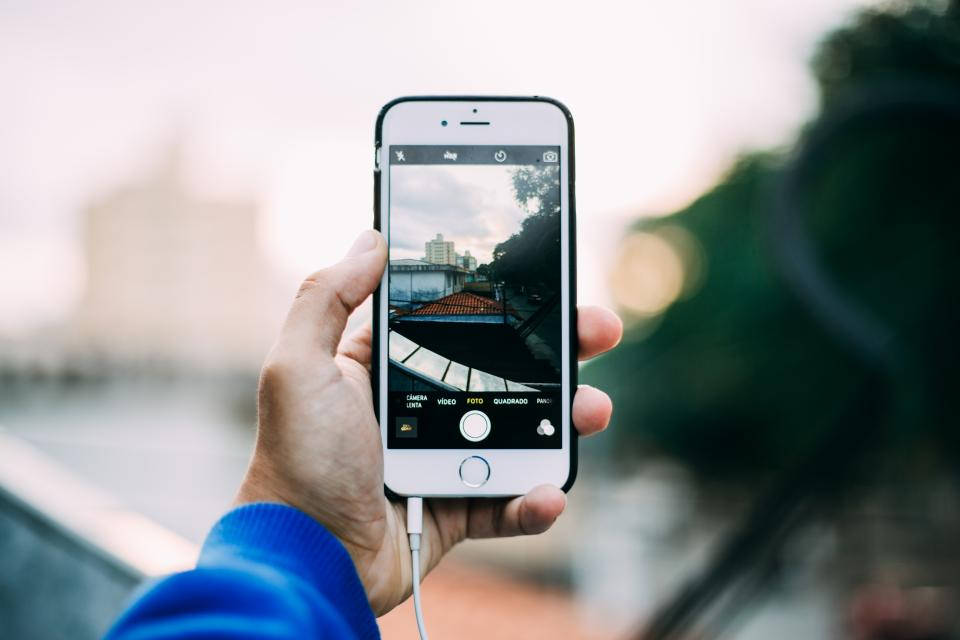 photography iPhone 桌布 壁紙 — 一隻手拿著智慧型手機，透過相機鏡頭捕捉模糊的城市景觀，包括屋頂和樹木。