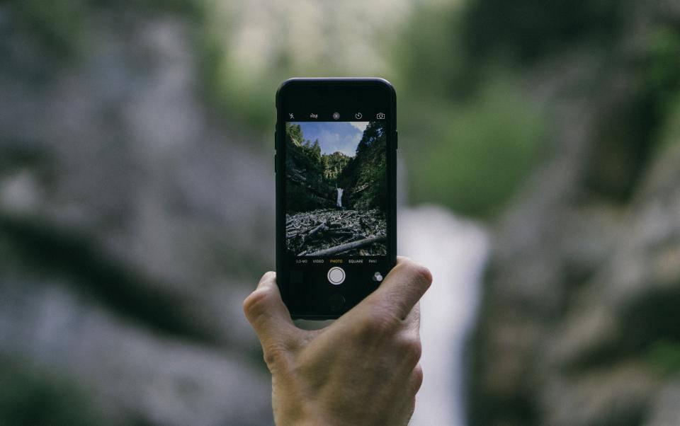 HD nature phone 桌布 壁紙 — 一隻手拿著智慧型手機，透過鏡頭捕捉茂密森林瀑布的場景。