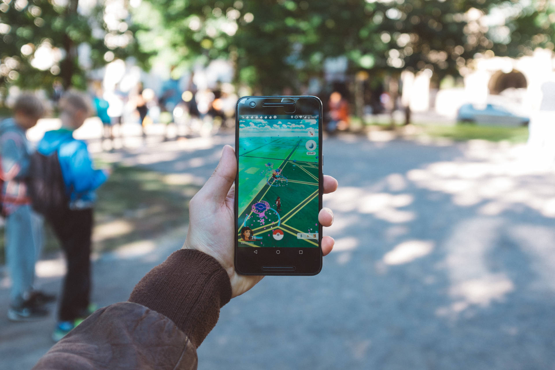 手拿著電話在陽光明媚的公園中玩 Pokémon Go，背景有人和樹木。 — 4K 寵物小精靈壁紙 壁紙